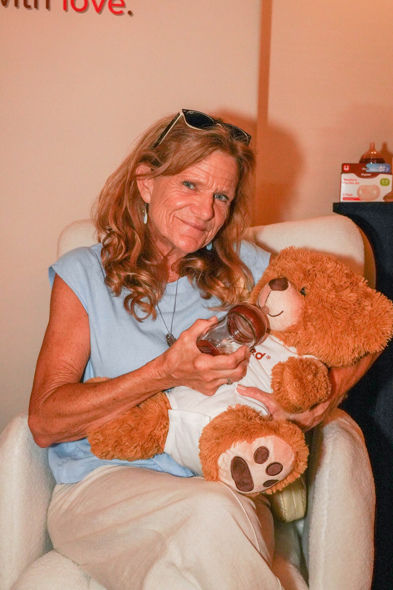 Woman with glasses holding a teddy bear, sitting on a chair with a large teddy bear, in a room with pink walls.