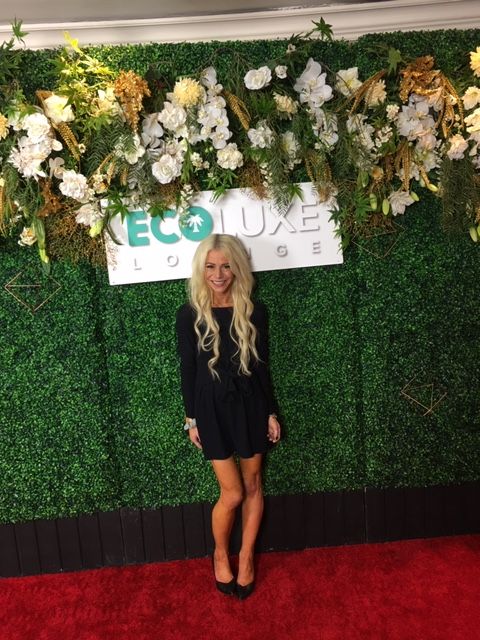 Woman with long blonde hair in black dress standing next to floral arrangement on green backdrop, red carpet below.