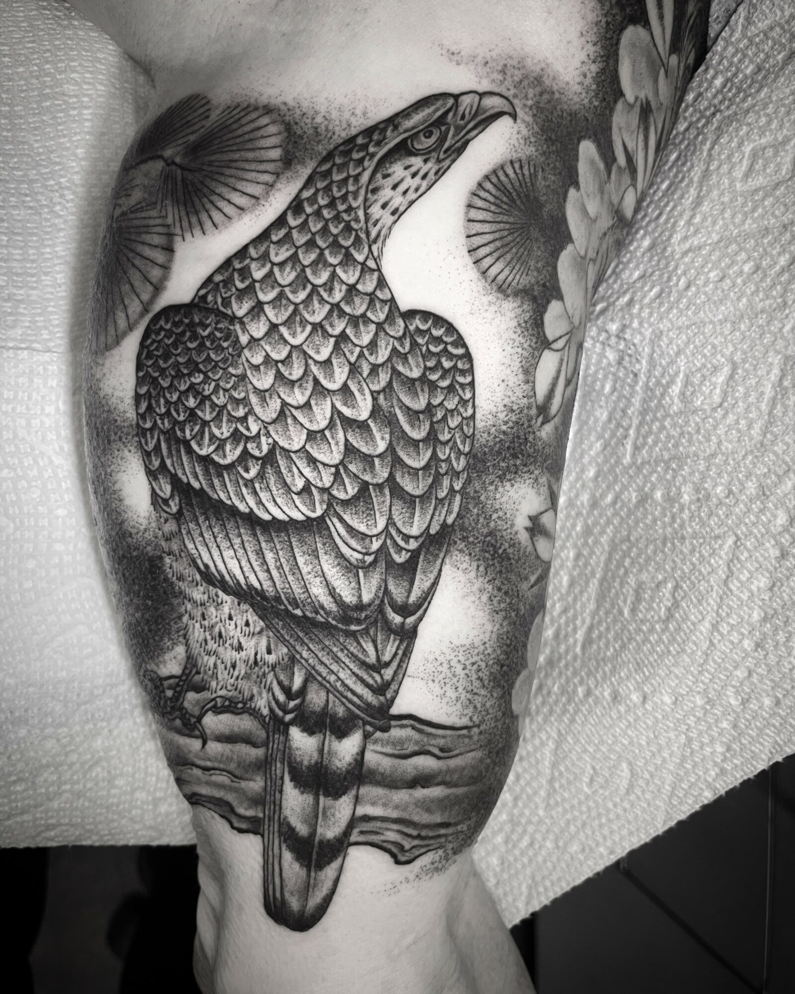 Black and gray tattoo of a bird with detailed feathers, surrounded by flowers and foliage, on a person's skin.