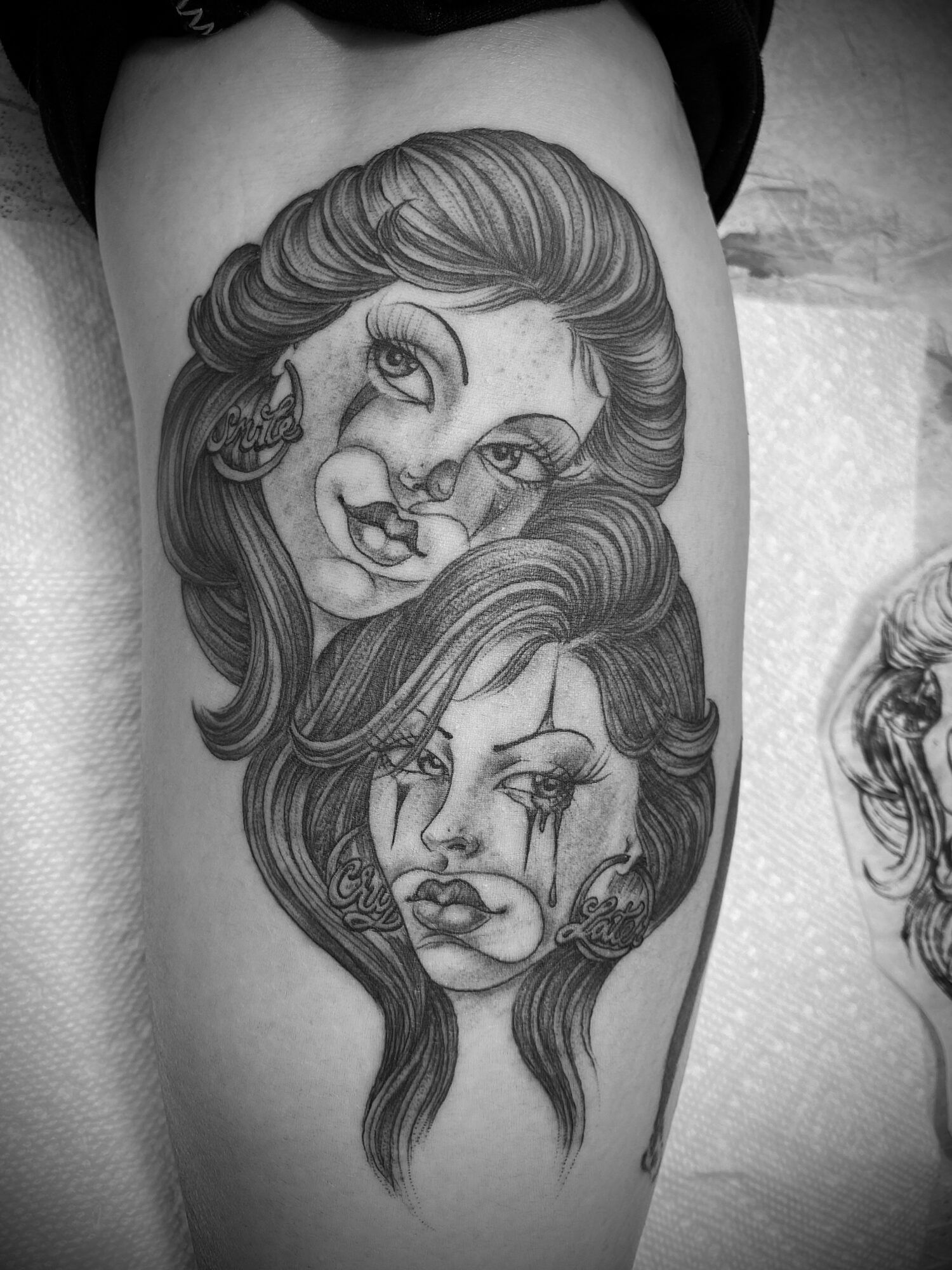 Black and white tattoo of two women's faces with detailed hair and expressive features on a person's thigh.