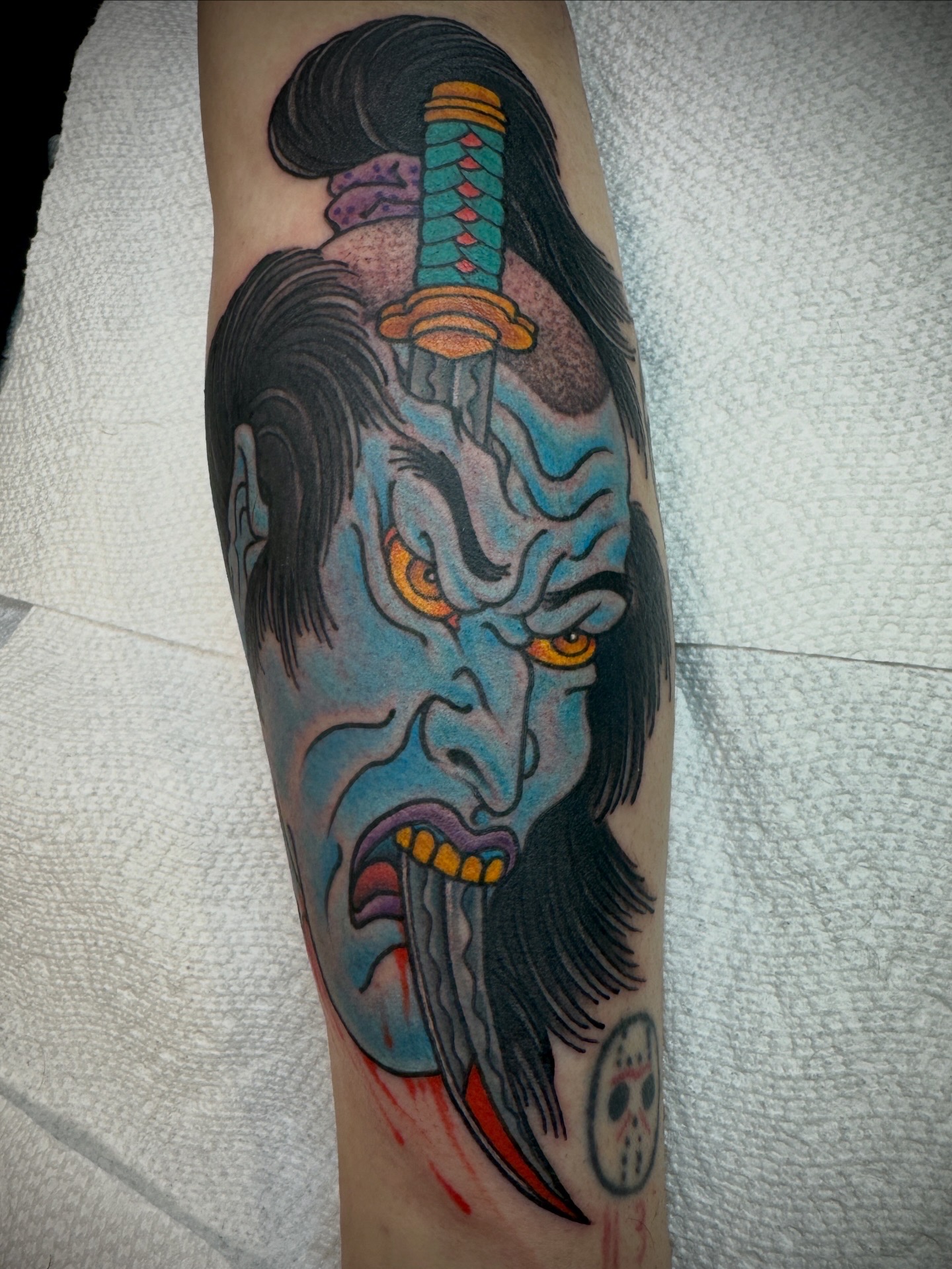 Tattoo of a blue-faced, fierce-looking demon with long black hair, yellow eyes, and a sword on the head, on a person's arm.