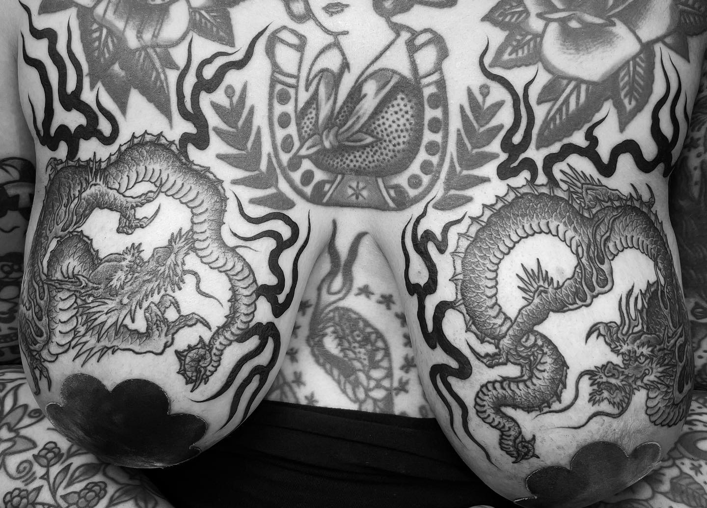 Black and white tattooed chest with two large dragons, floral patterns, and a central figure with a bird and decorative elements.