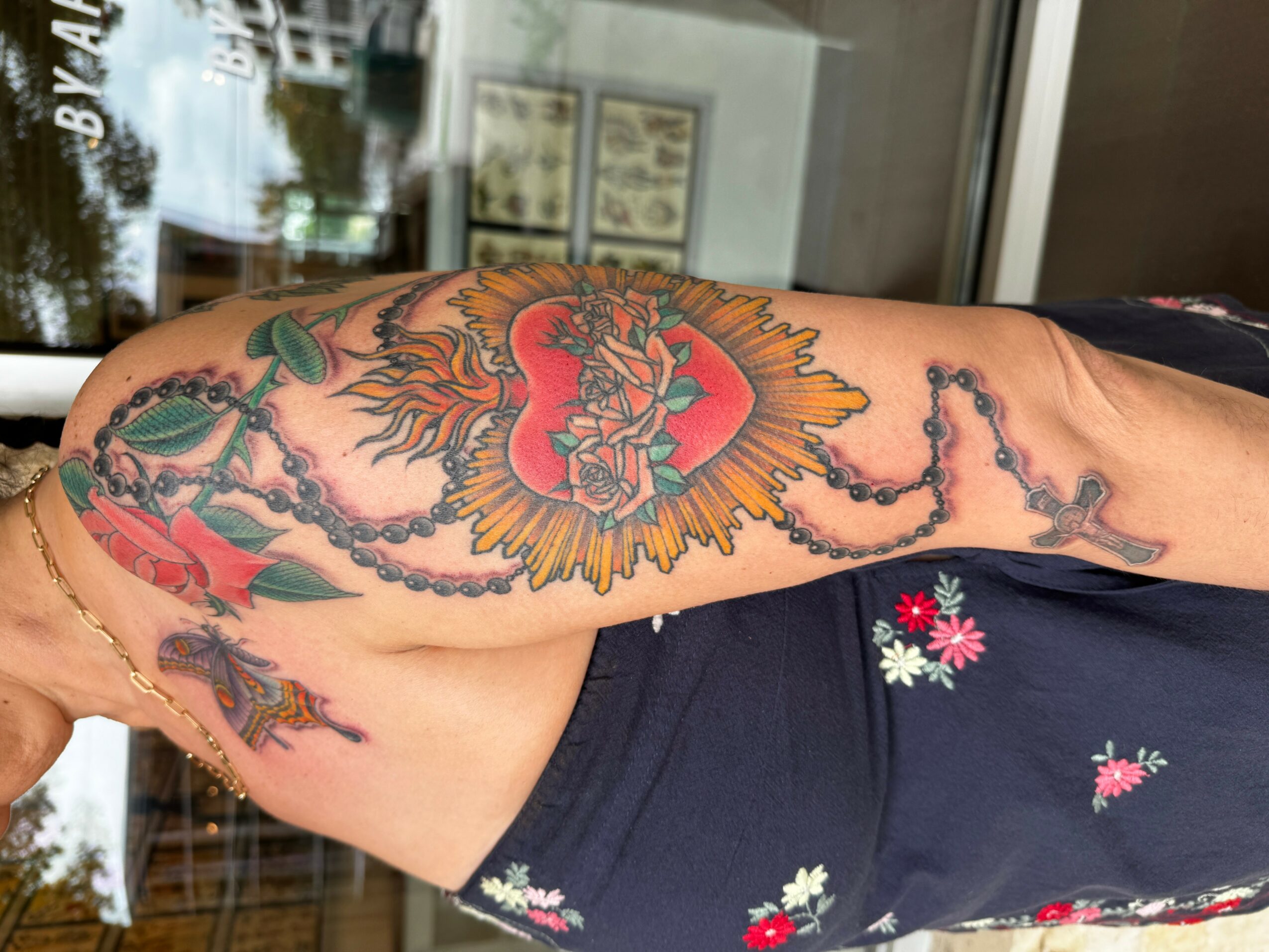 Tattoo of a lion's face inside a heart with wings on upper arm, surrounded by a rosary, colorful and detailed.
