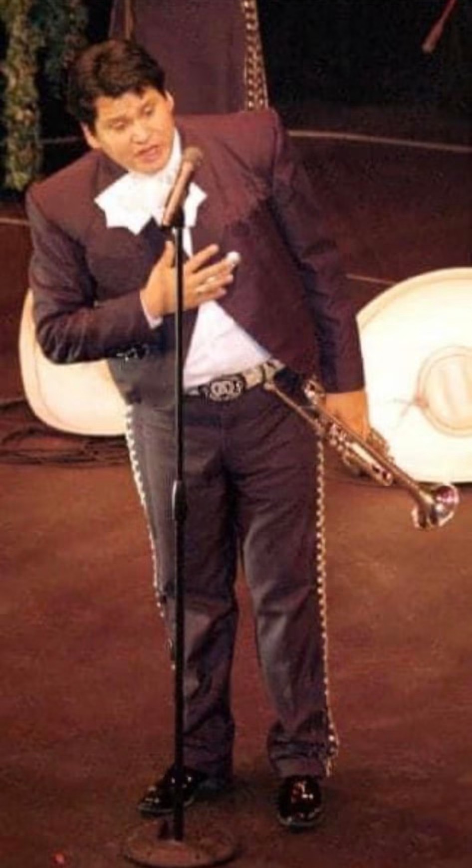Person in formal attire holding a microphone and a cane on stage, with a white hat nearby.