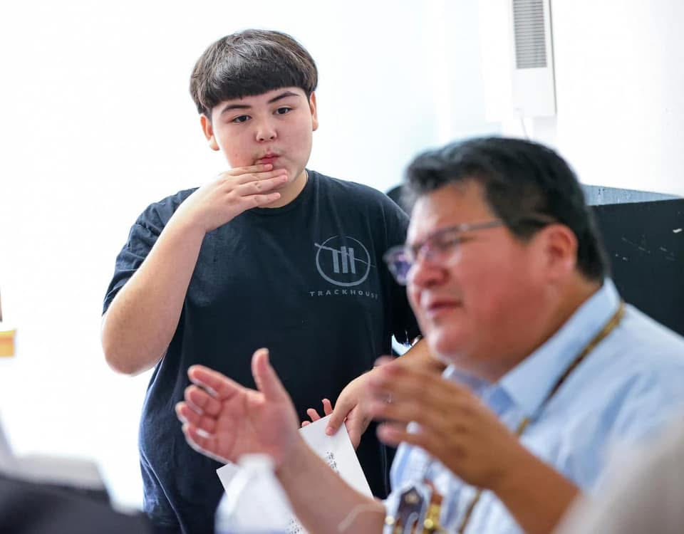 Boy and adult in conversation indoors, boy touching chin, adult gesturing with hands, bright background, casual setting.