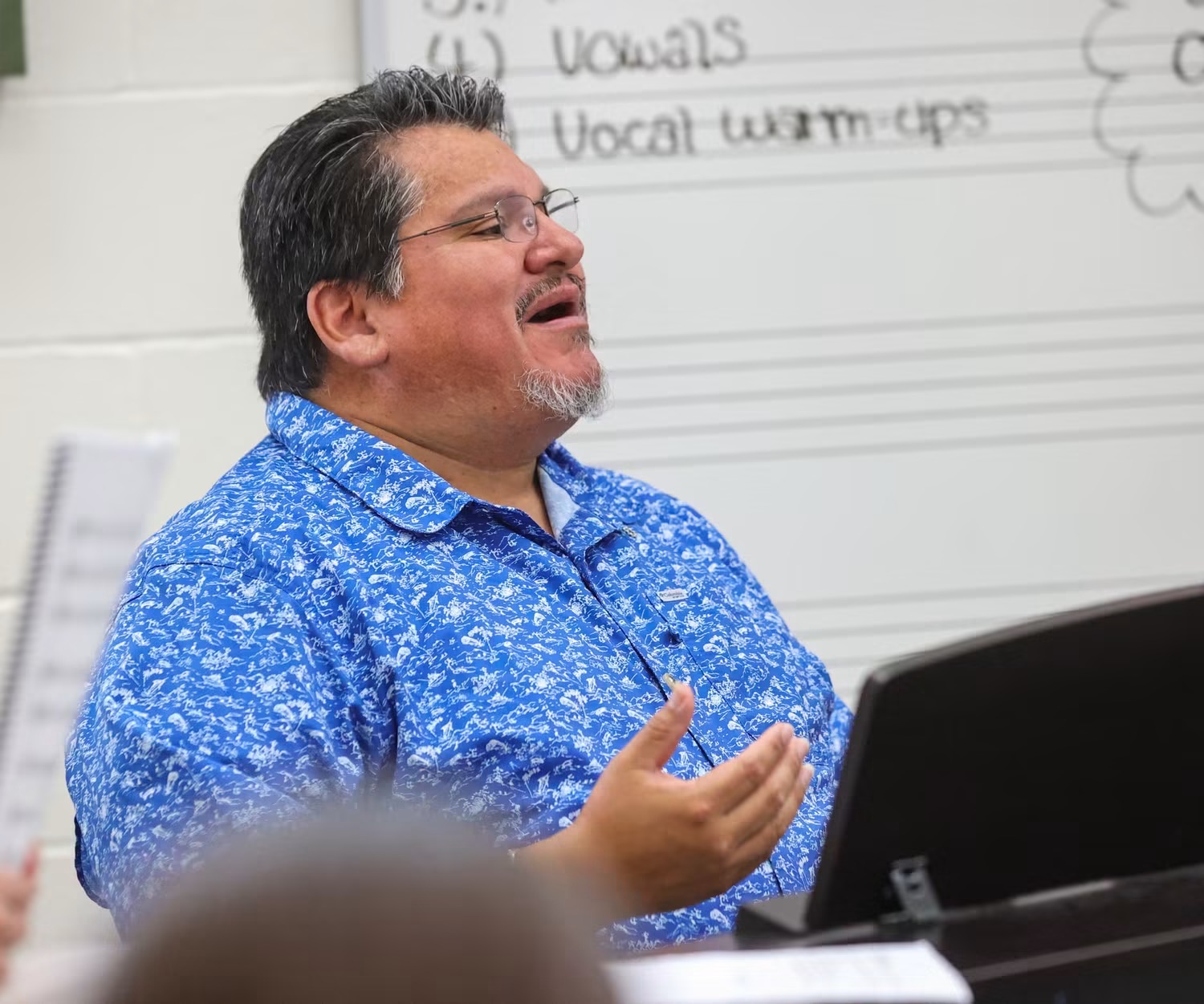 Man with glasses and gray hair, wearing a blue patterned shirt, speaking in front of a whiteboard with writing.