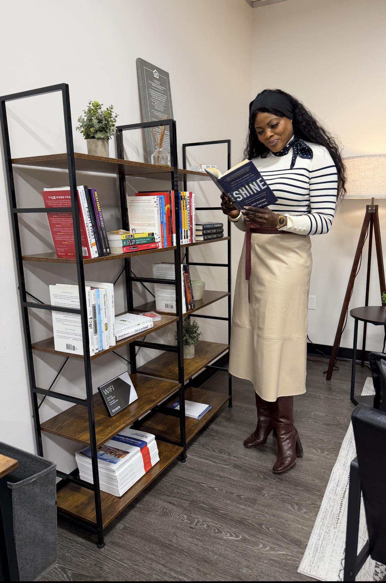 Woman reading a book in a room with a black metal and wood bookshelf, white walls, and a wooden floor.