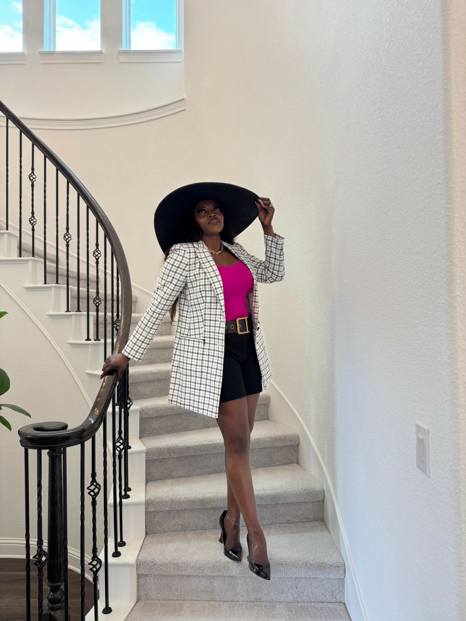 Woman in a checkered blazer, pink top, black shorts, and high heels standing on staircase with large hat, indoors.