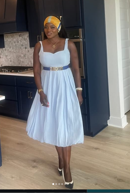 Woman in white dress with yellow headscarf standing in kitchen with dark cabinets and light wood floor.