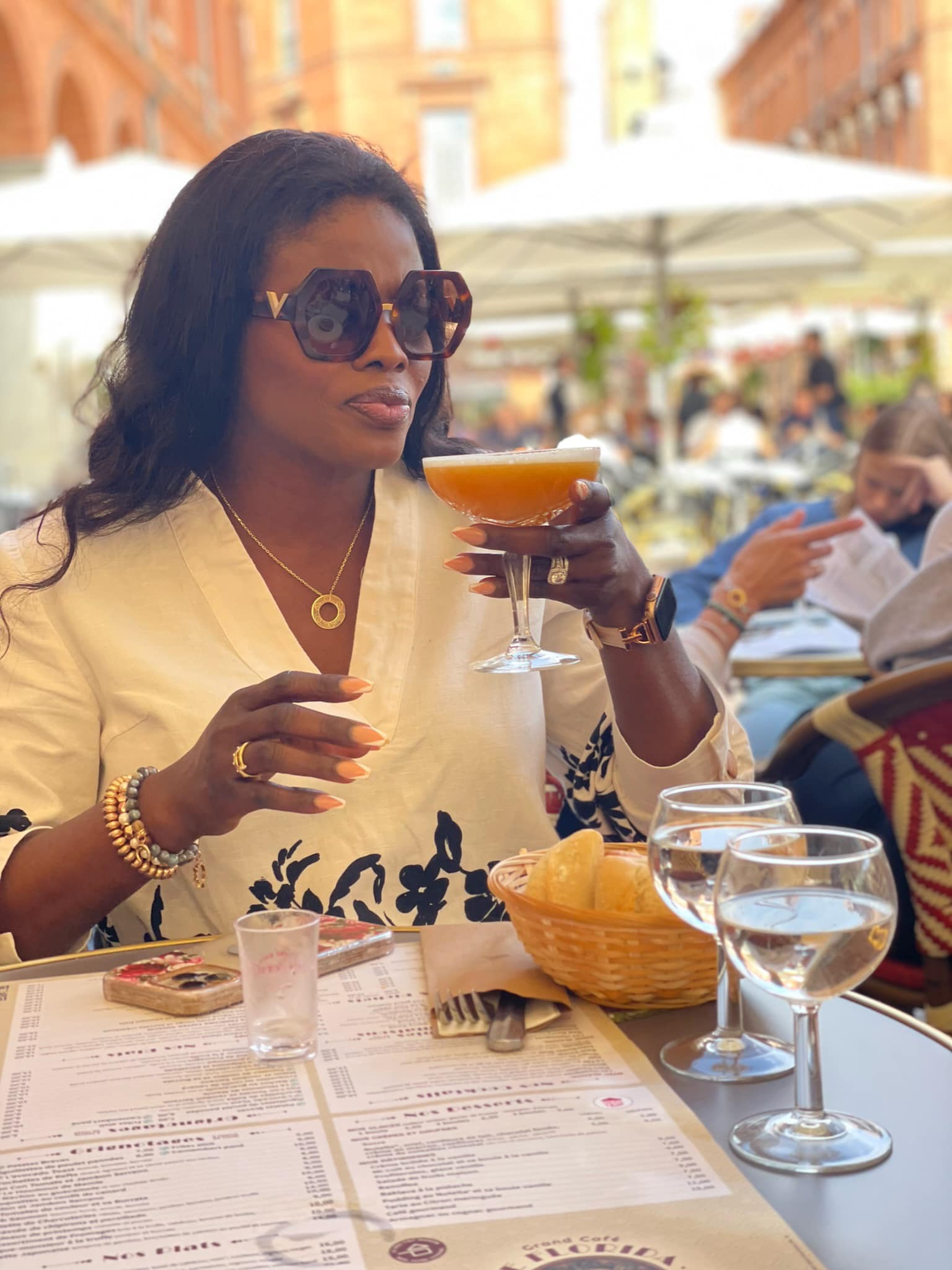 Woman with dark hair and sunglasses holding a cocktail at an outdoor restaurant, with glasses and a basket on table.