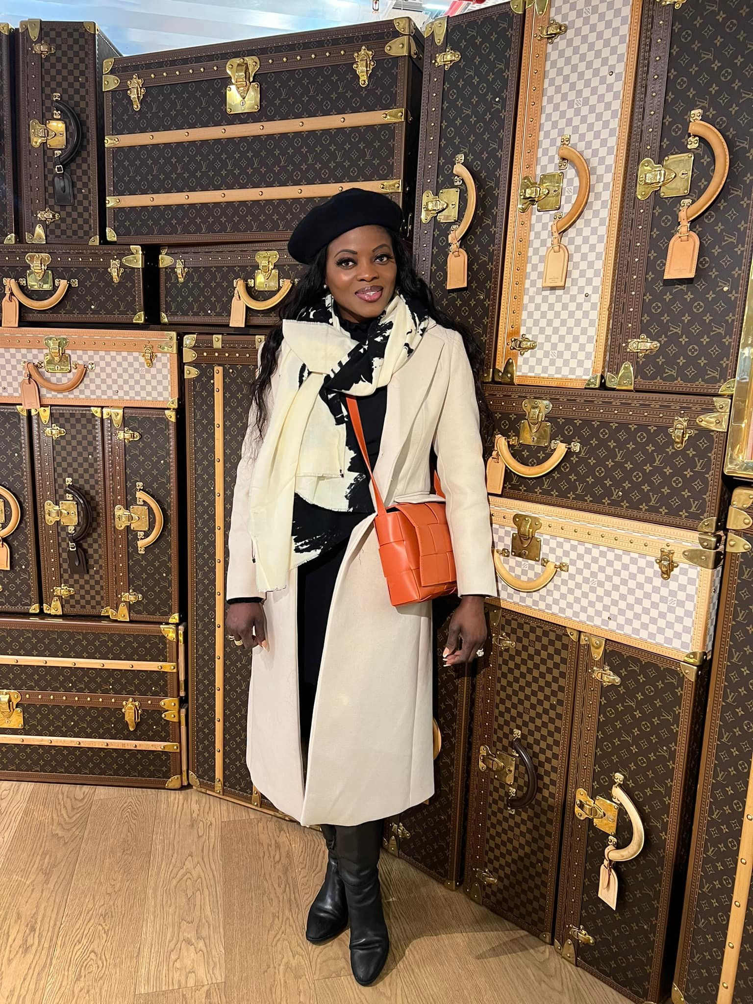 Woman in a white coat and black boots standing among stacked patterned suitcases, smiling, with a black beret and orange bag.
