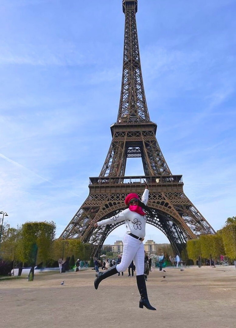 Person jumping in front of the Eiffel Tower with a pink hat and white outfit, blue sky background.