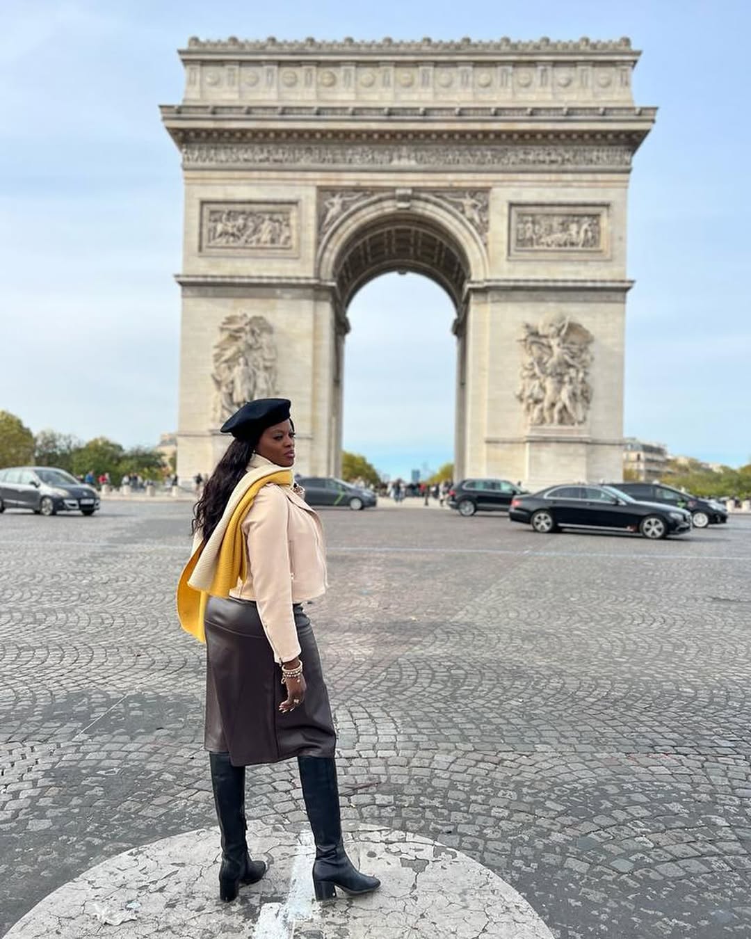 Woman standing on cobblestone street in front of Arc de Triomphe, facing left, with cars in background.