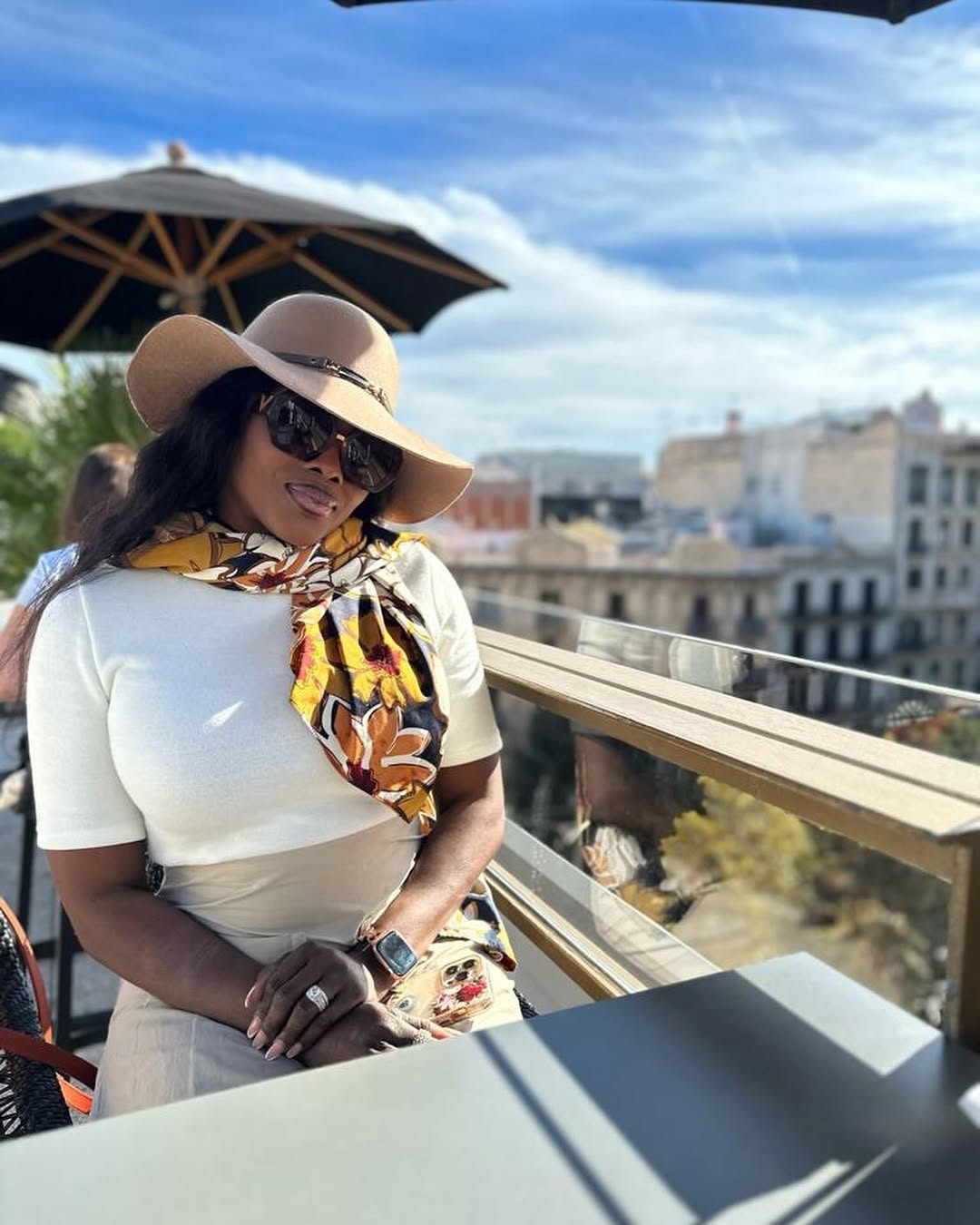 Woman wearing a wide-brimmed hat, sunglasses, and a scarf, sitting outdoors with a cityscape background under a blue sky.