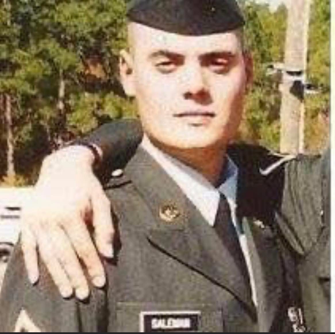 Young man in military uniform with a cap, outdoors with trees in background, looking at camera, serious expression.
