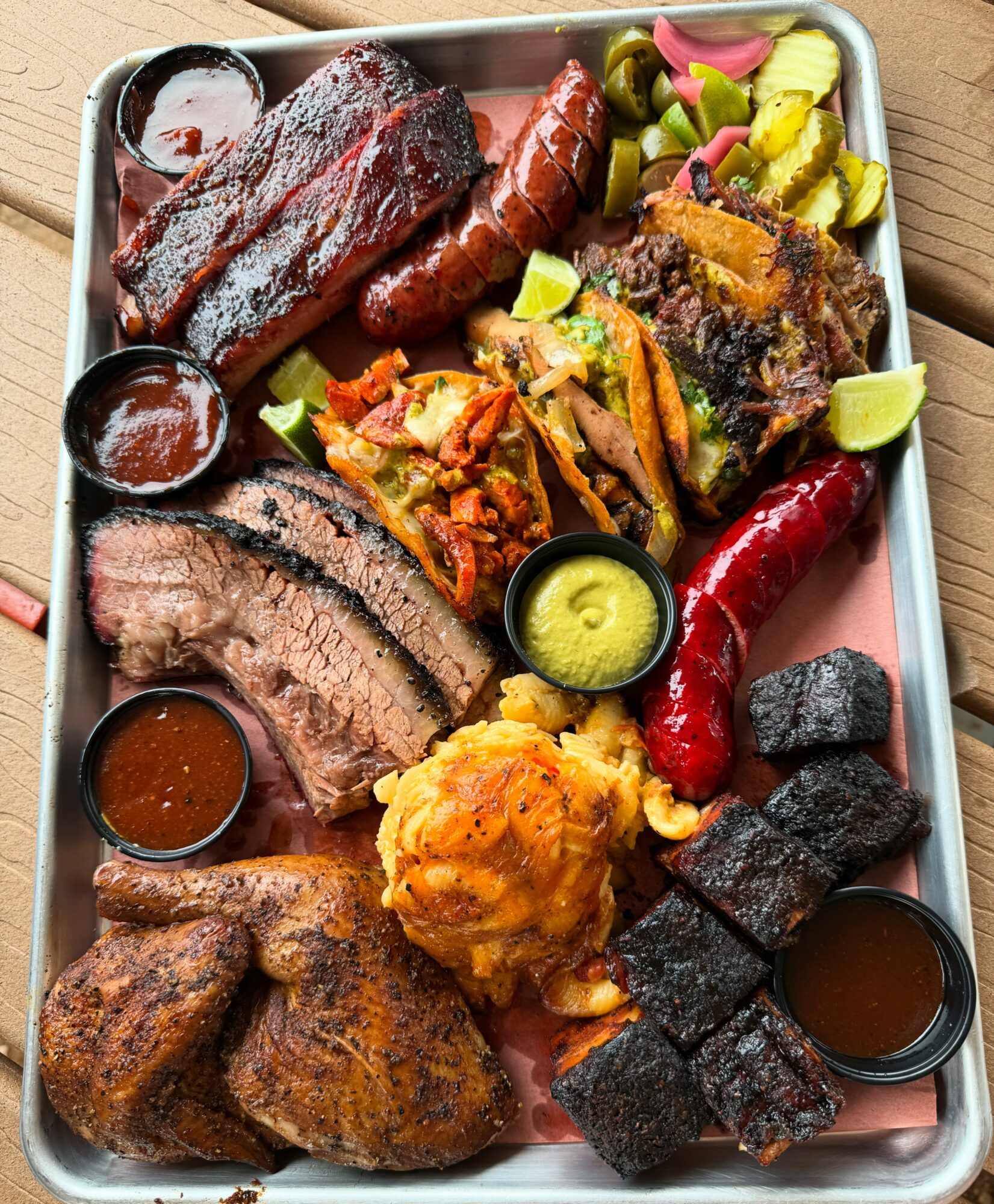 Assorted barbecue meats, sides, and sauces on a tray, including ribs, sausage, pulled pork, fried chicken, and vegetables.