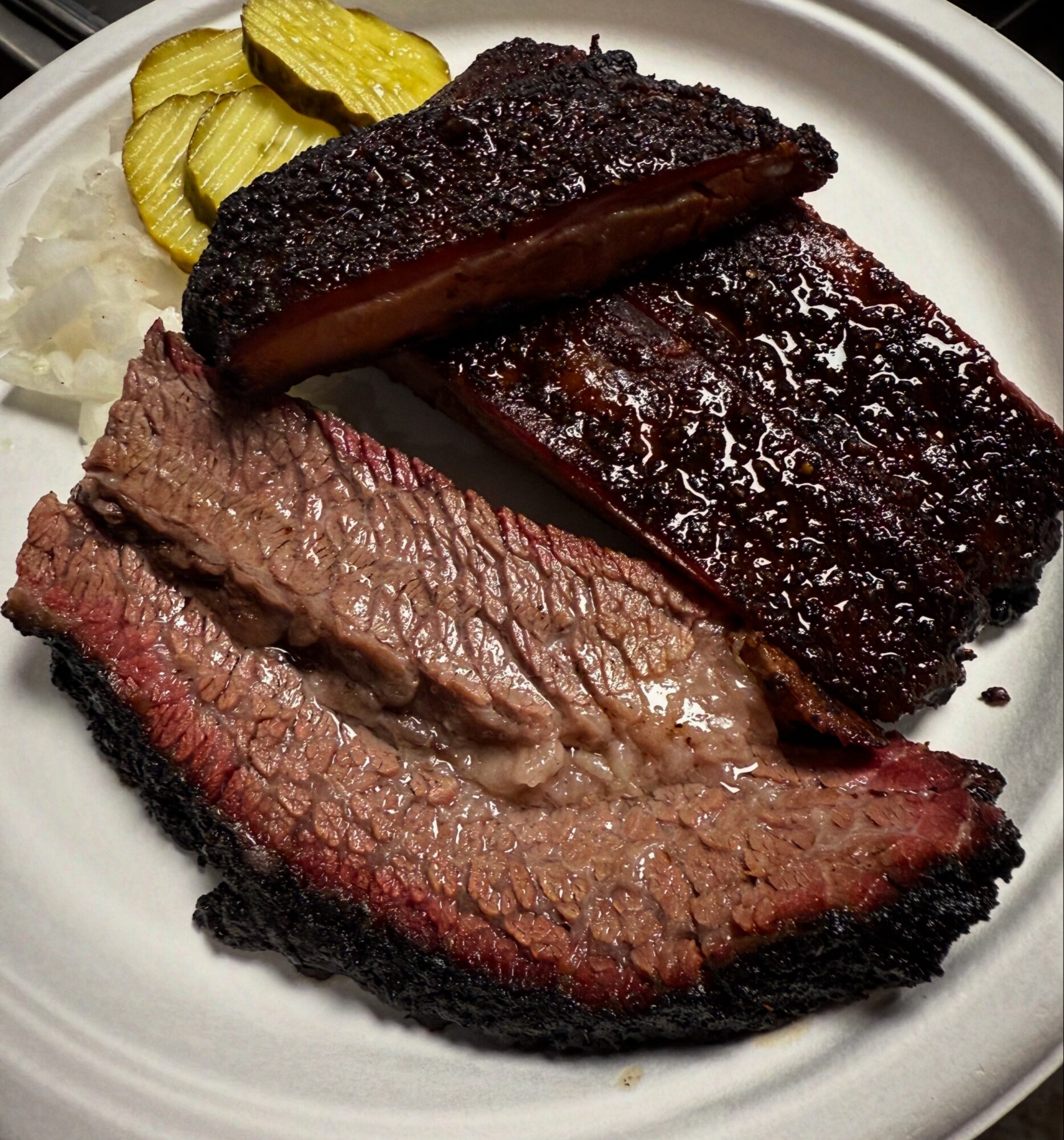 Plate with sliced cooked steak, barbecue ribs, pickles, and onions, all on a white dish.