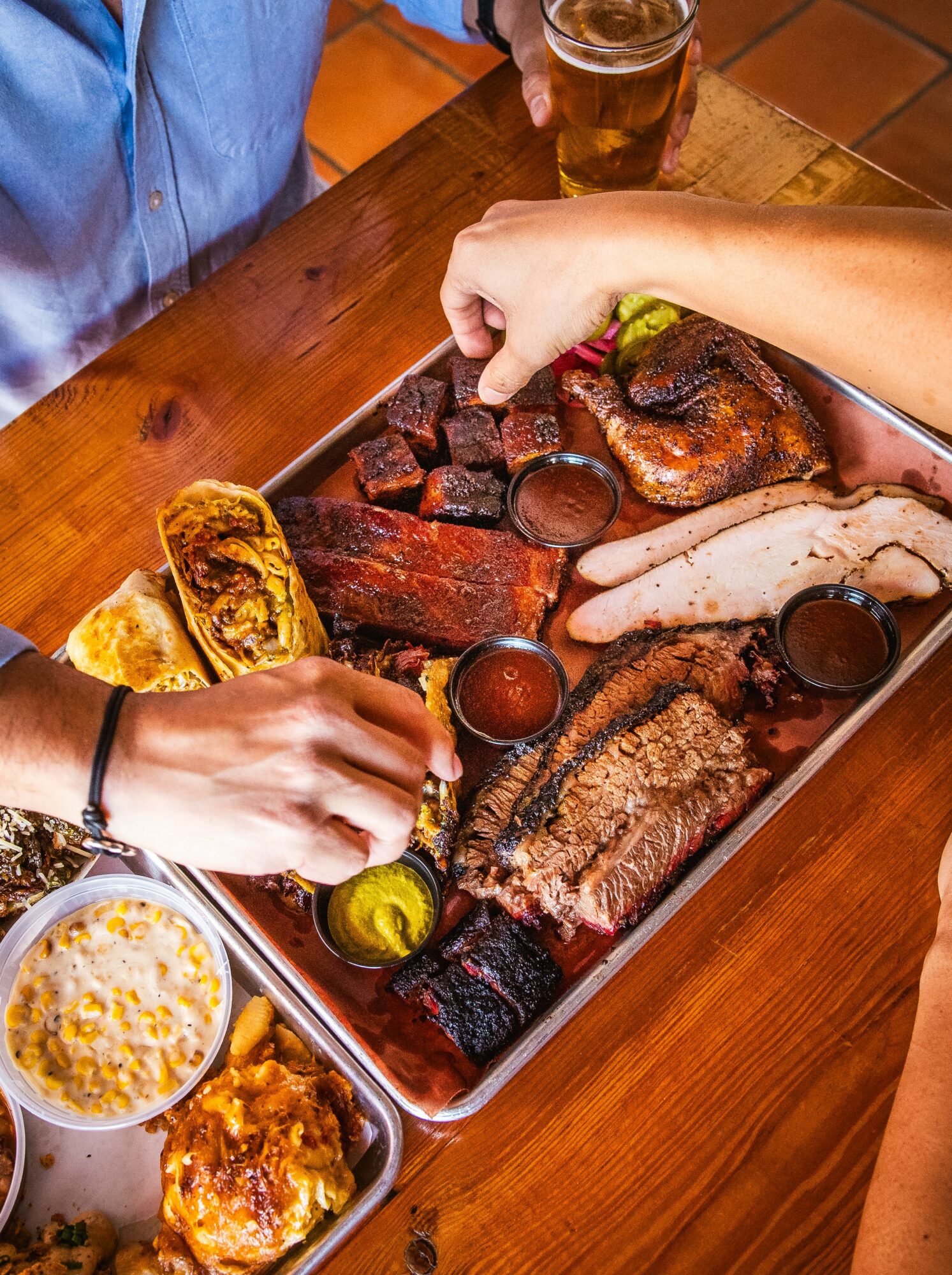 People sharing a tray of grilled meats, sauces, and bread at a wooden table with drinks, hands reaching for food.