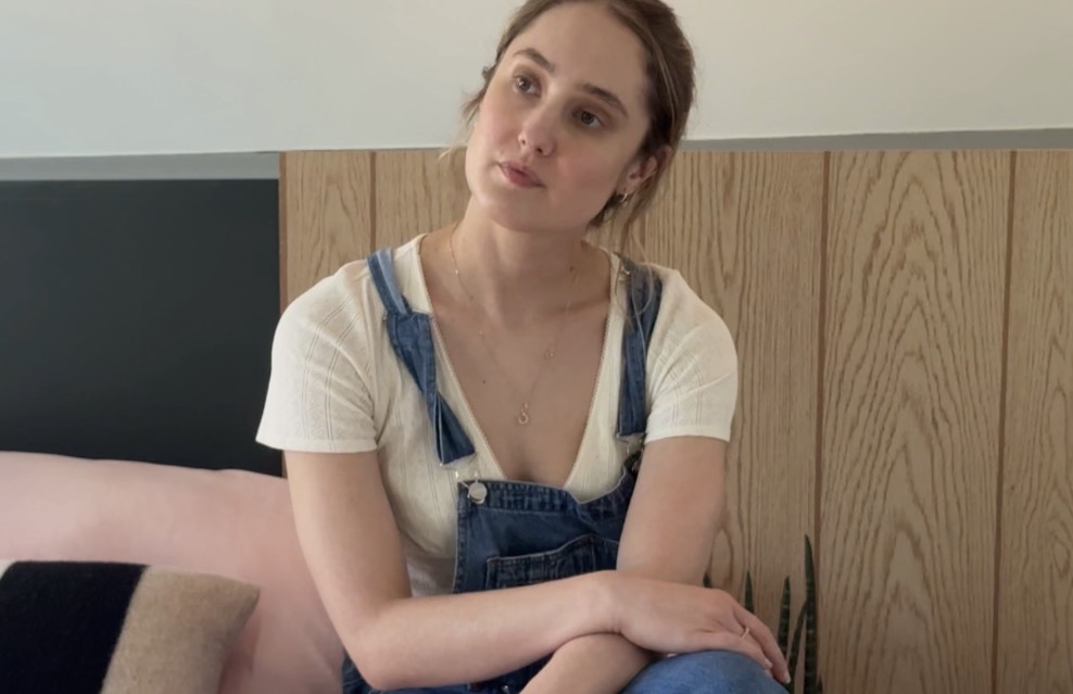 Young woman with brown hair sitting on a bed, wearing a white shirt and denim overalls, looking to the side.