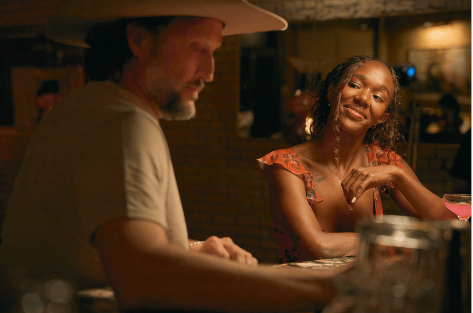 A man and woman sit at a bar, smiling and engaging in conversation, with warm lighting in the background.