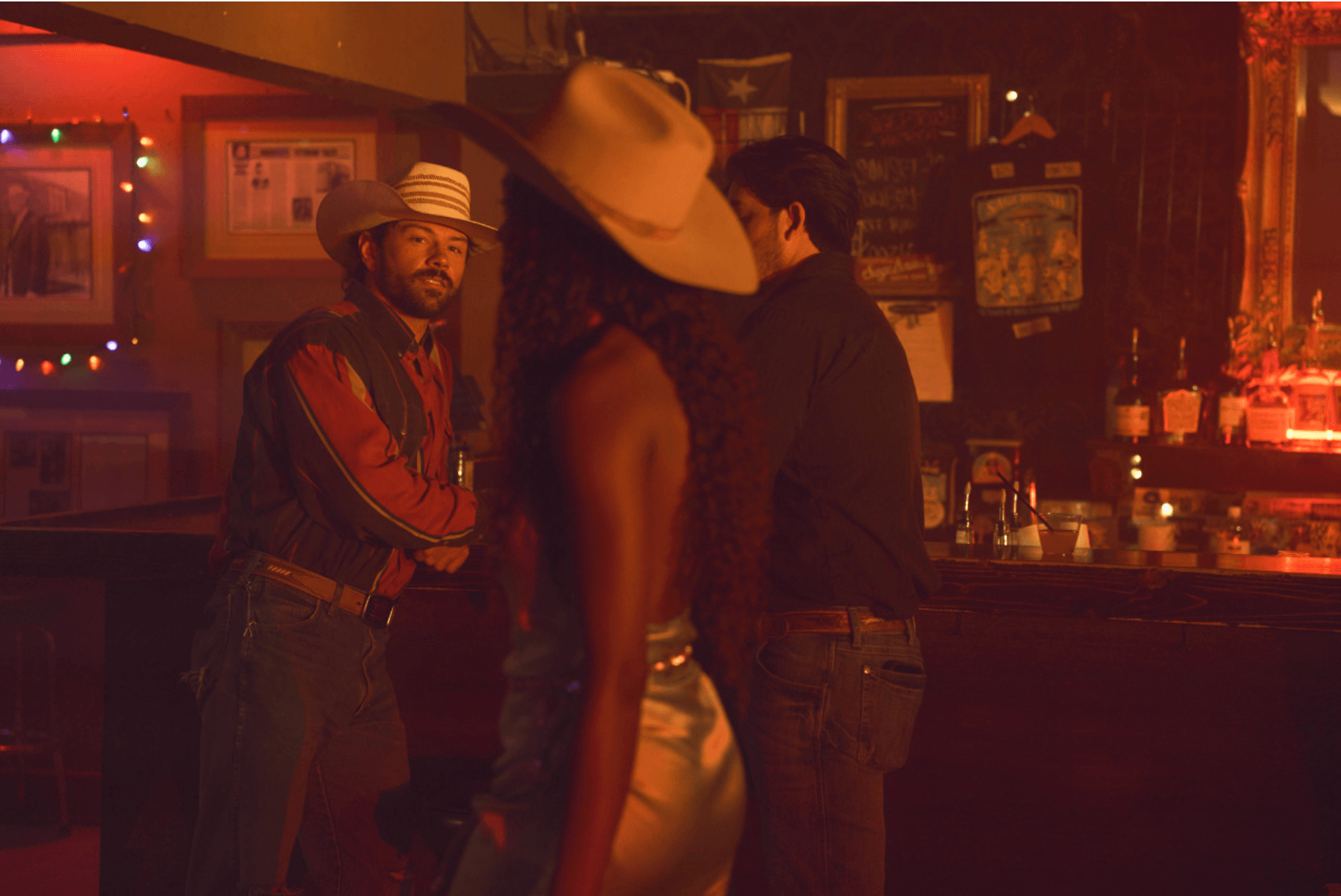 Three people in a dimly lit bar, one wearing a large hat, two facing each other, one smiling, warm lighting.