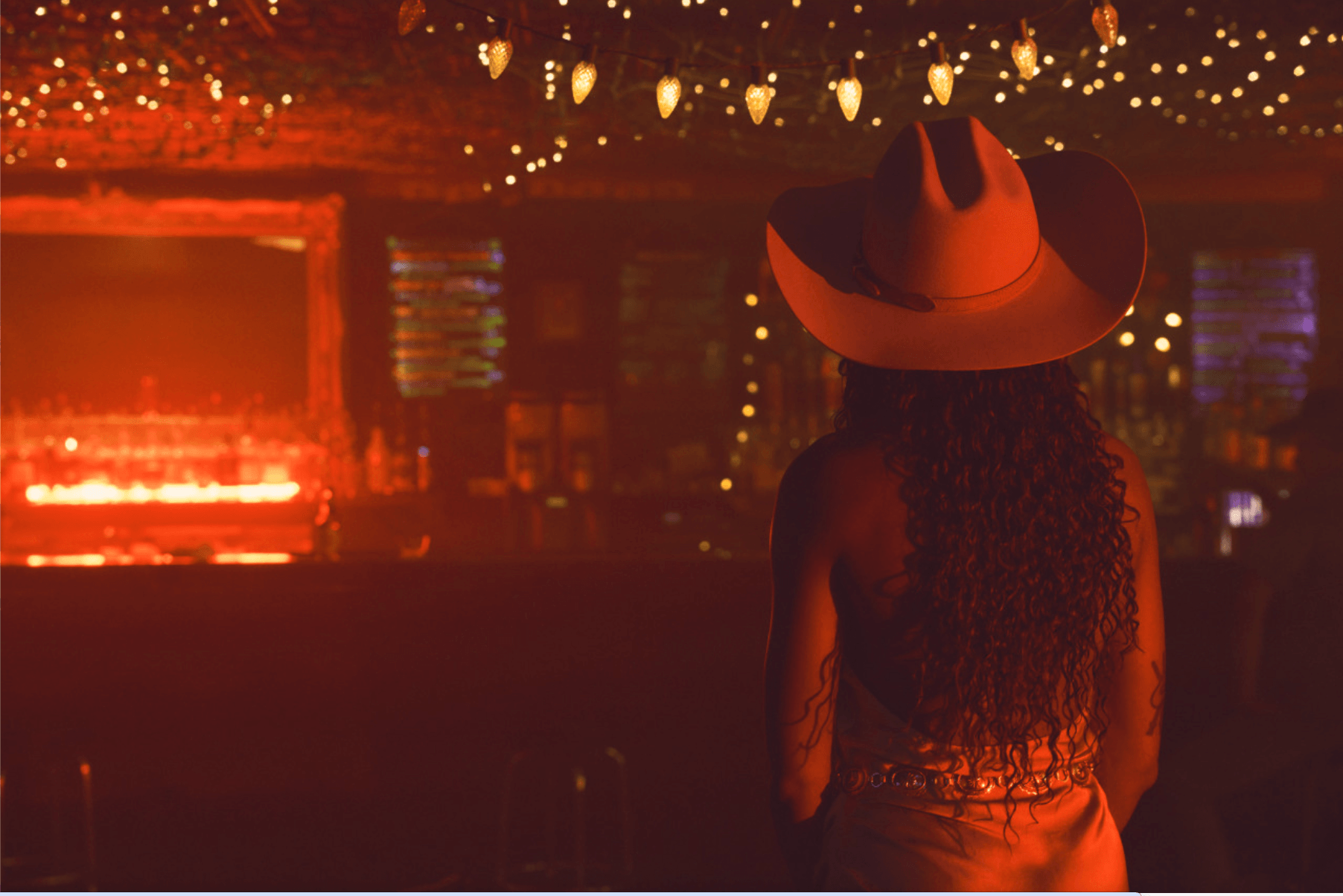Person wearing a large hat with a wide brim, facing away in a dimly lit indoor space with string lights.