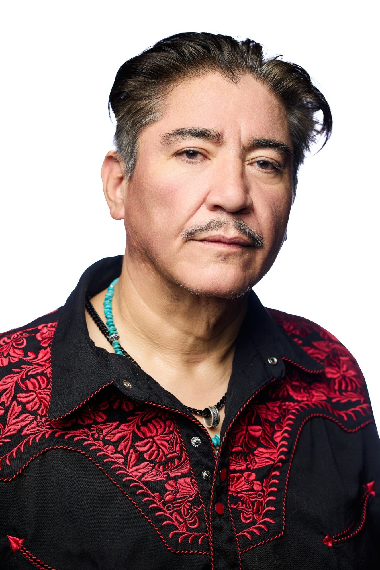 Portrait of a middle-aged man with dark hair, wearing a black and red embroidered jacket and a turquoise necklace.