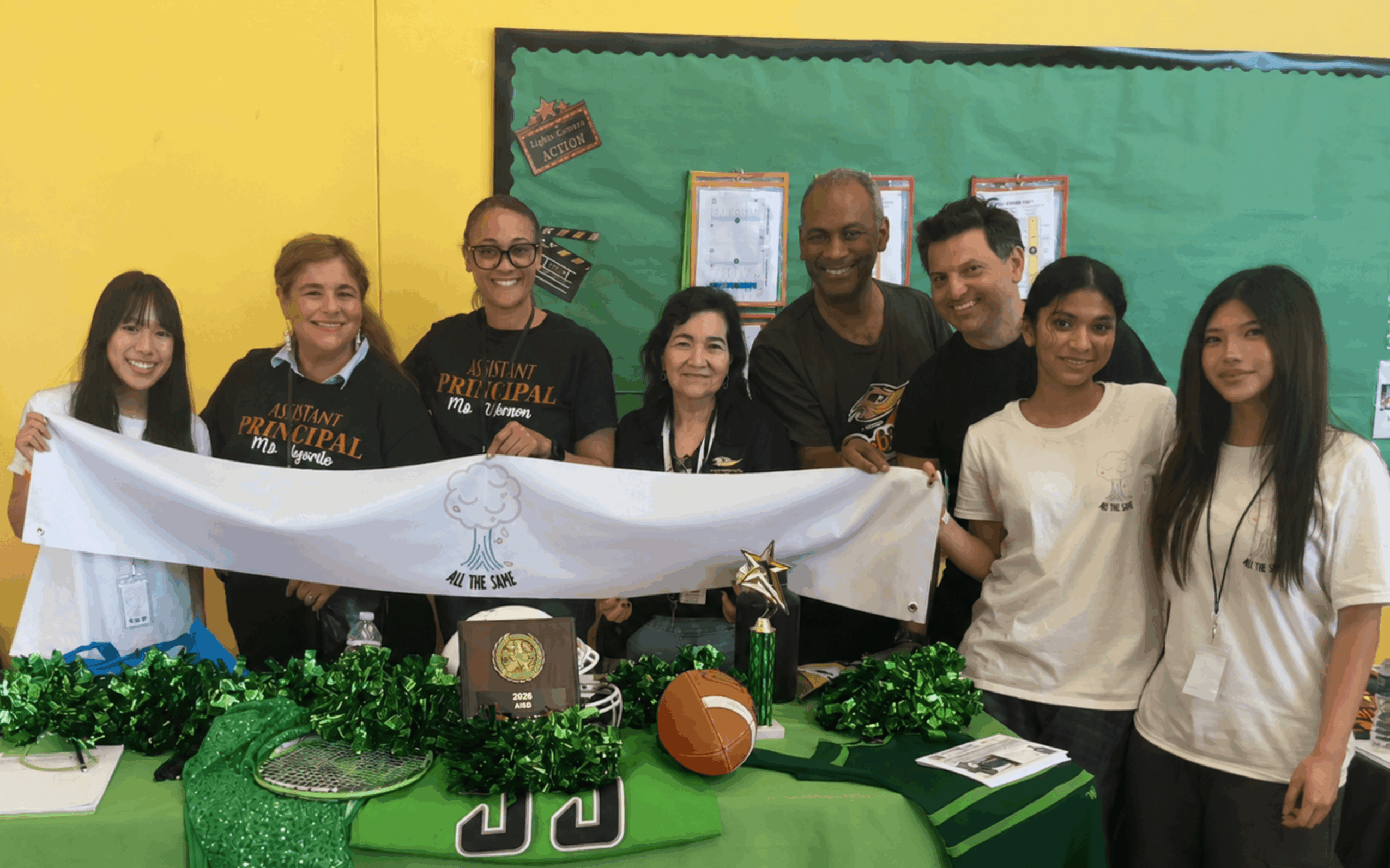 Group of eight people standing behind a table with sports-themed decorations and a banner, in a classroom setting.