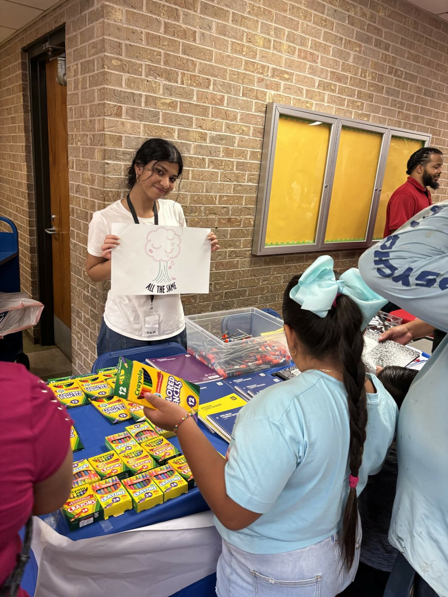 Girl holding a drawing at a table with snacks, others around, brick wall background, window, and people in the scene.