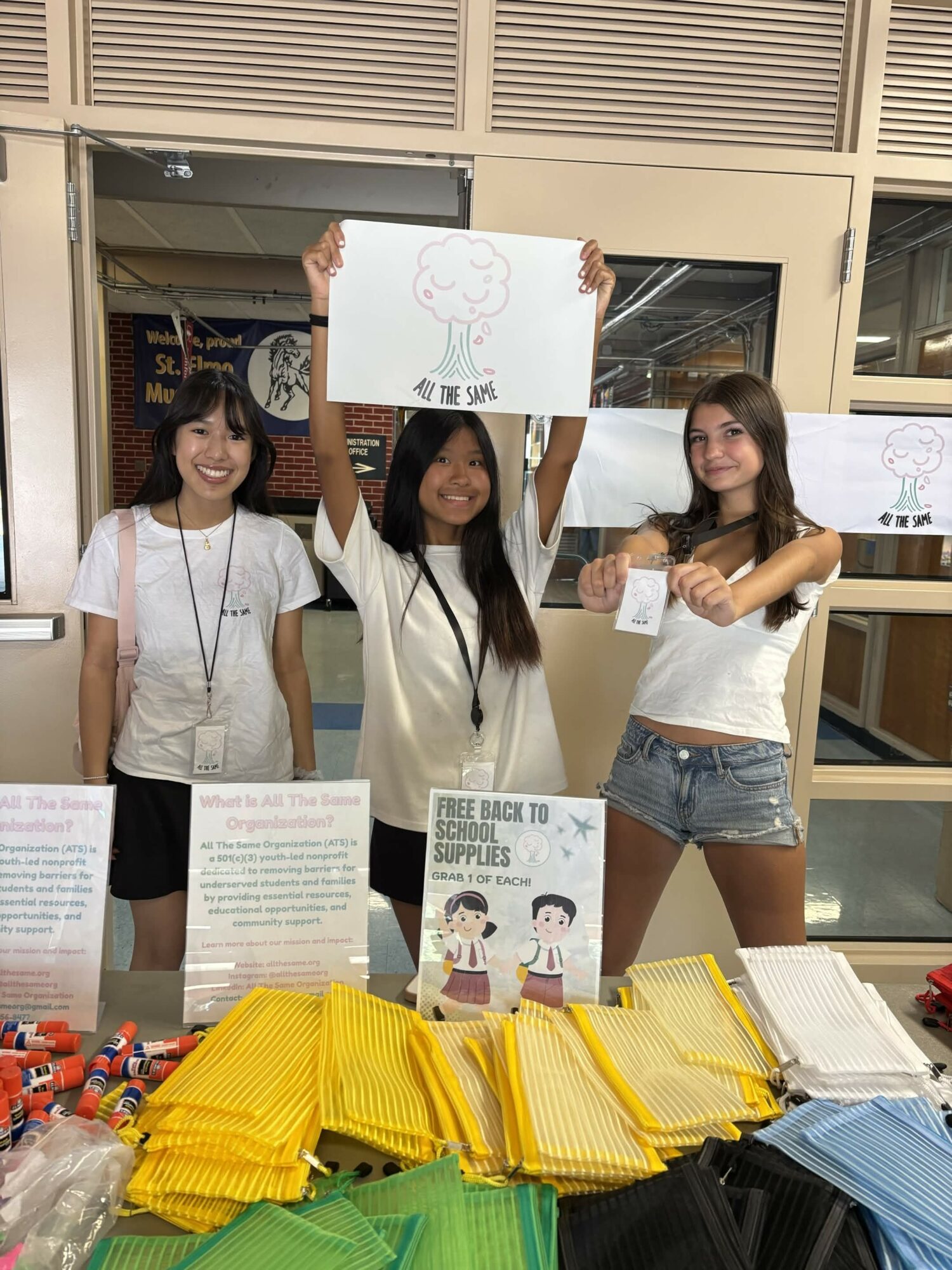 Three young women standing behind a table with yellow and blue bags, holding a sign with a tree drawing, smiling.