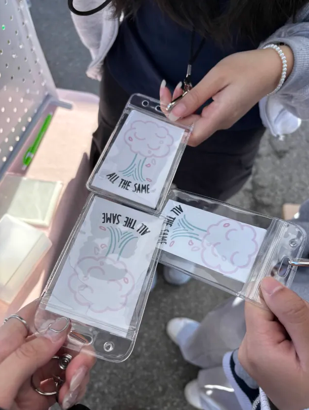 Two people hold transparent plastic cases with cloud illustrations and text, one person wears a bracelet, background shows a gray surface.