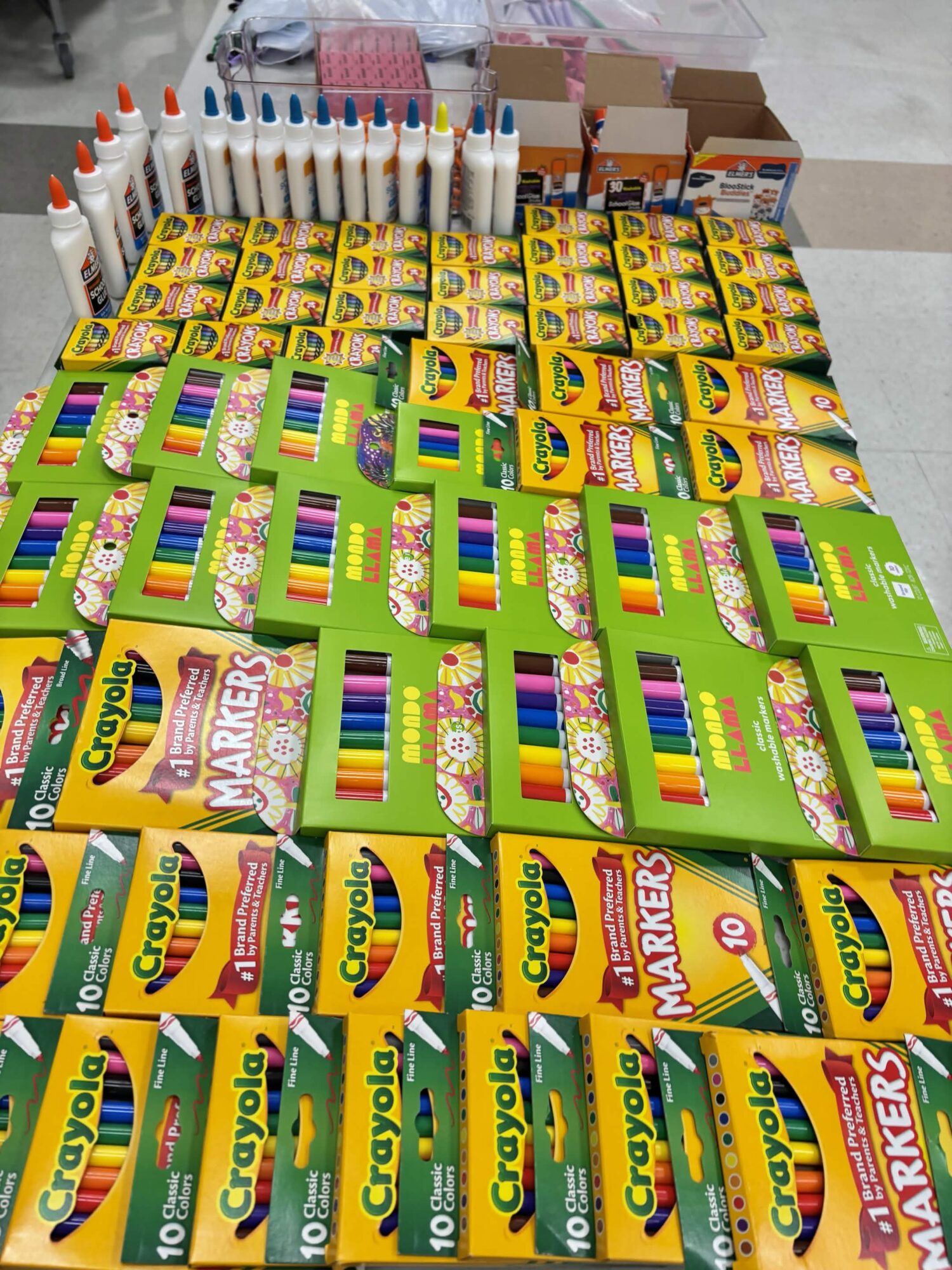 Table with Crayola markers, colored pencils, and art supplies arranged in rows, with boxes and bottles in the background.