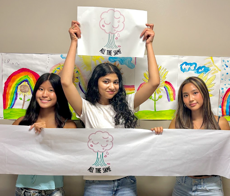 Three young women holding a banner with a cloud and rainbow drawing, standing in front of colorful rainbow and cloud artwork.