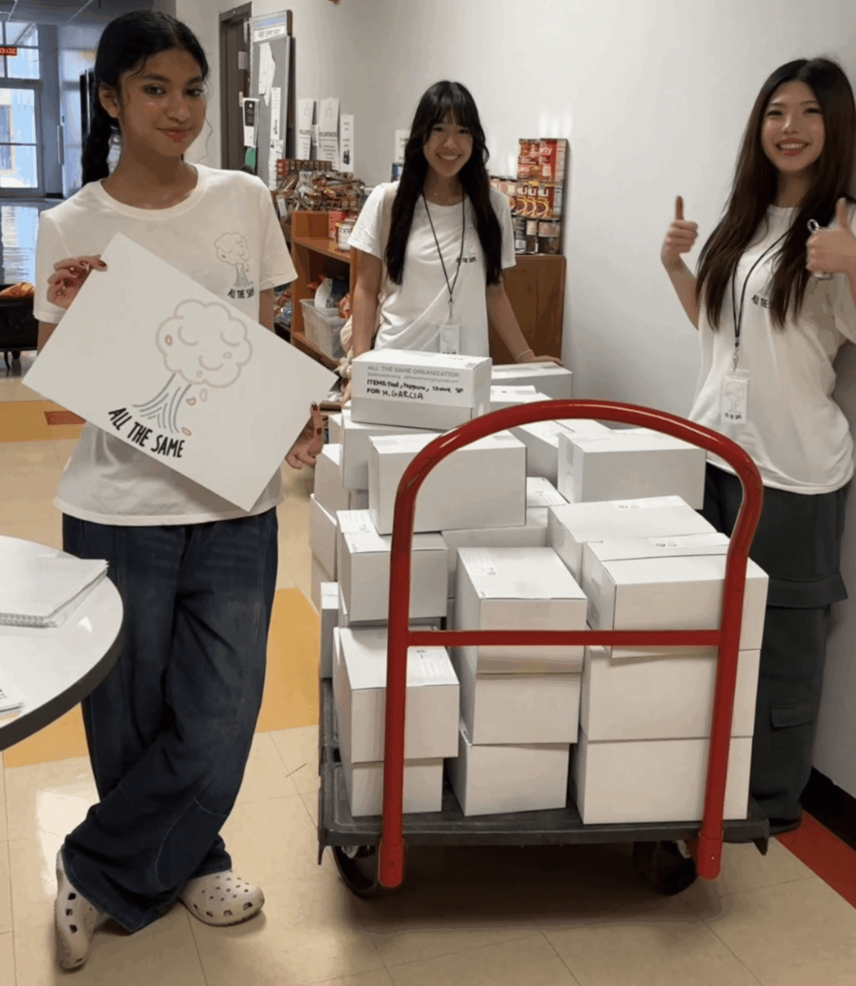 Three women standing behind a cart filled with white boxes, smiling and giving thumbs up, in a room with shelves and boxes.