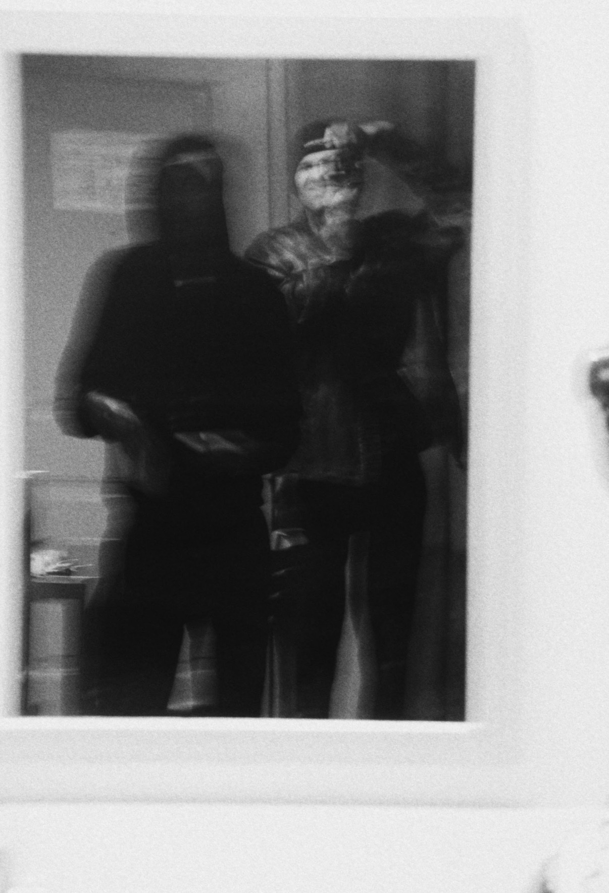 Two people taking a mirror selfie, one holding a phone, in a room with a door and some objects visible.