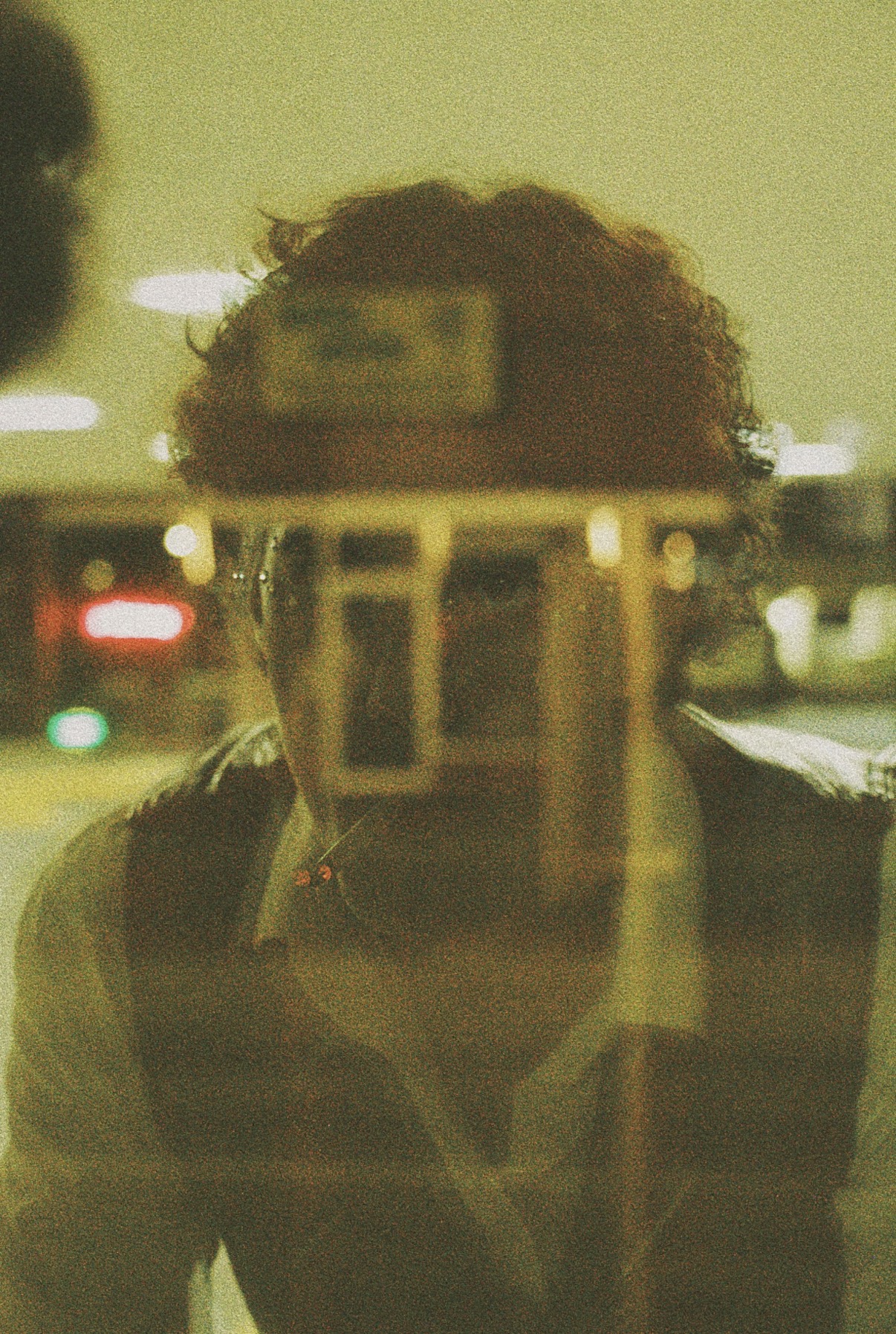 Person with curly hair taking a photo in a mirror at night, city lights visible behind.