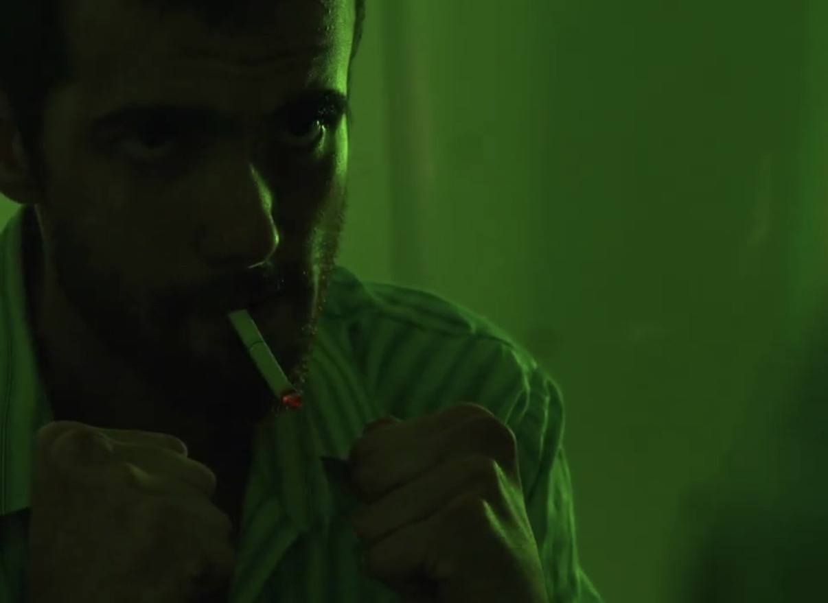 Person with a beard and short hair, holding a cigarette, in a dimly lit green environment.