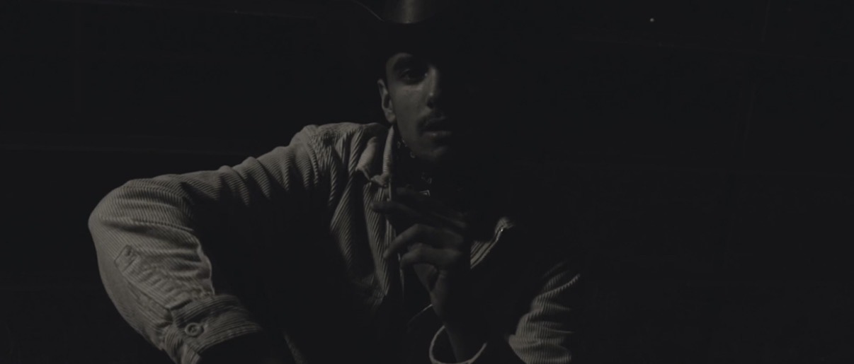 Person sitting in a dark environment with minimal lighting, face partially visible, wearing a jacket.