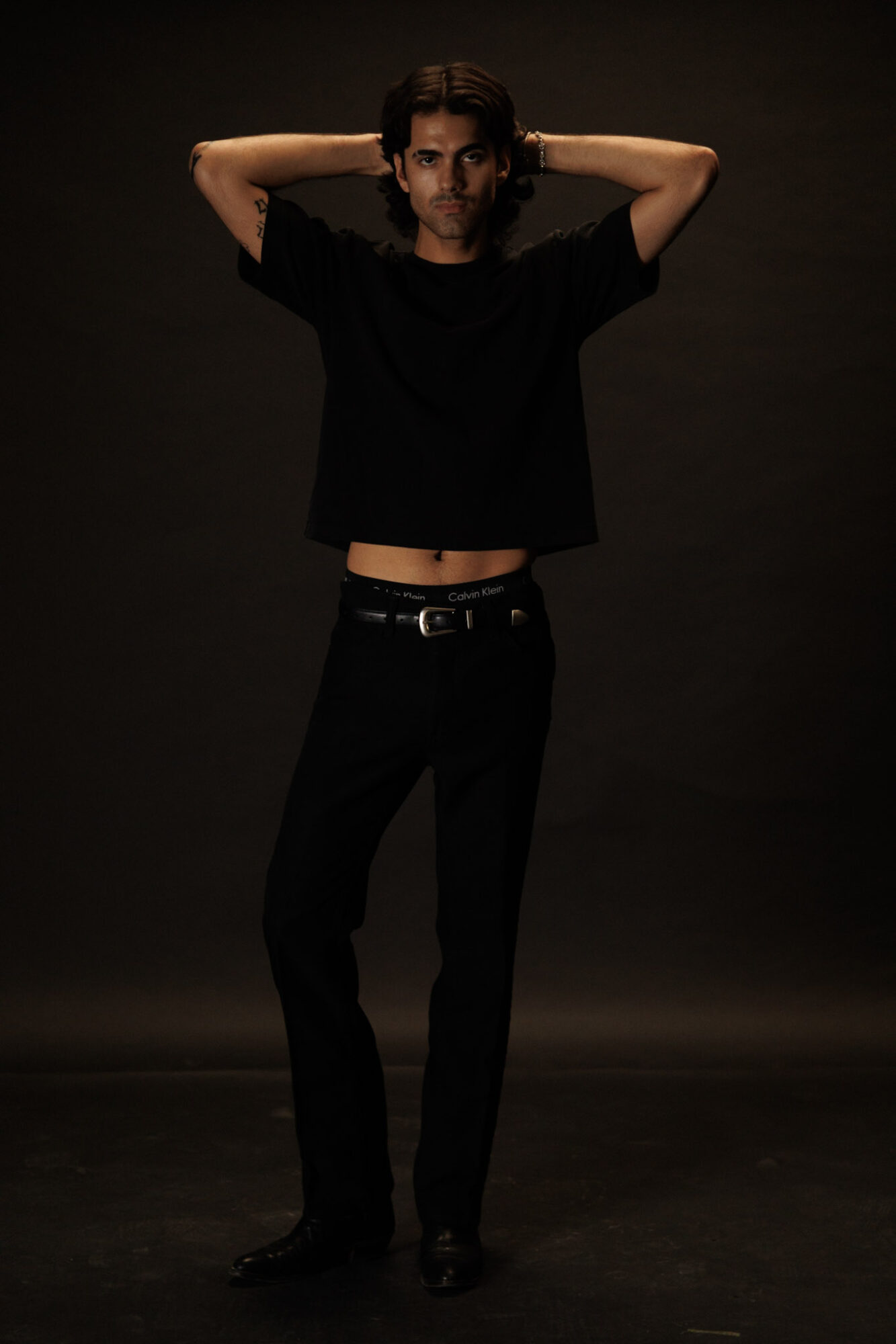Person standing with arms behind head, wearing black shirt and pants, against dark background.