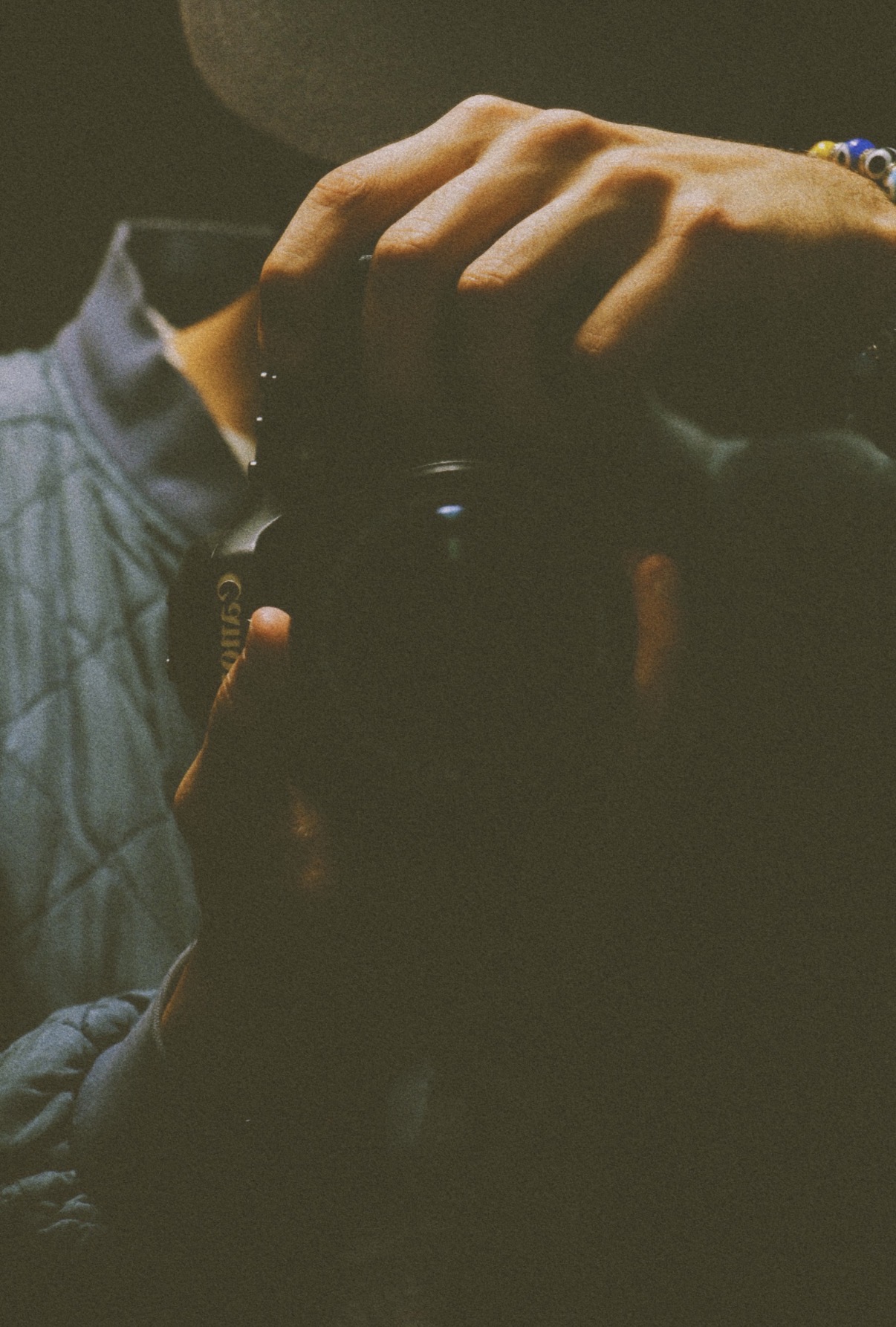 Person holding a camera, hand resting on head, dark background, dim lighting, wearing a jacket.