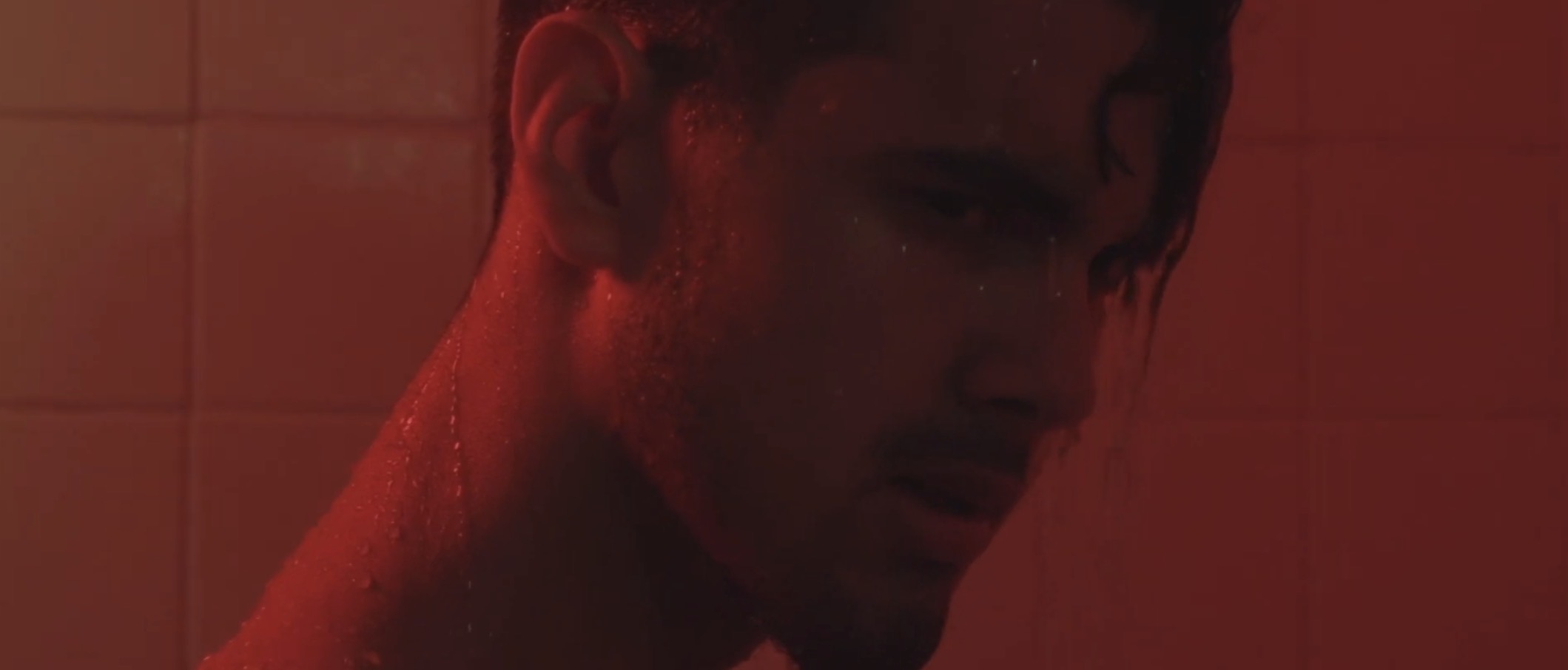 A young man with wet hair in a dimly lit room with red lighting, looking downwards.