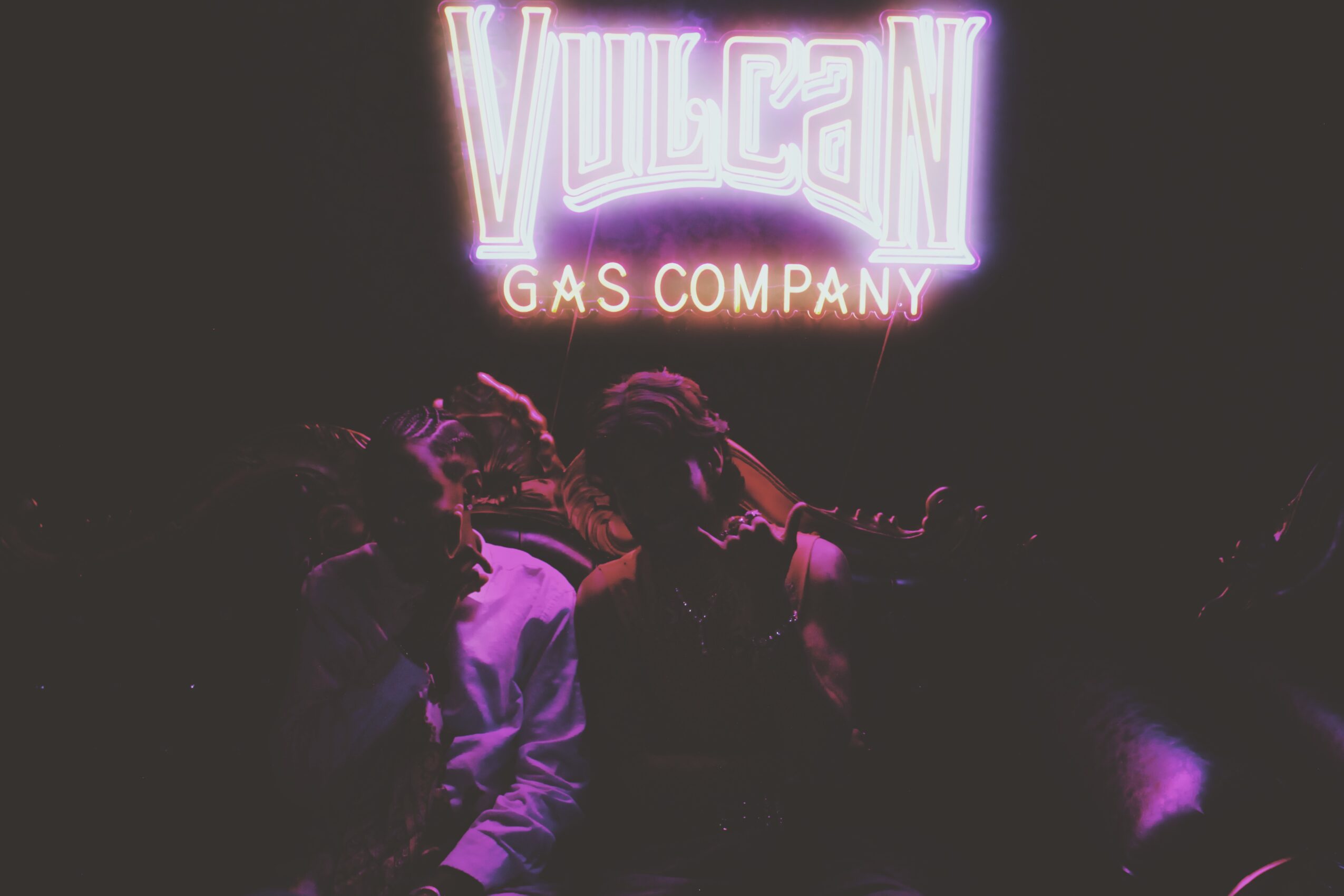 People in front of a neon sign reading 'Vulcan Gas Company' at night, with dark surroundings and purple lighting.