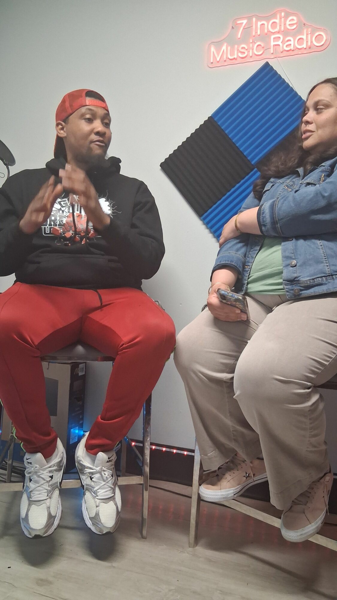 Two people sitting on chairs, one holding a phone, in a room with a sign and soundproofing panels on the wall.