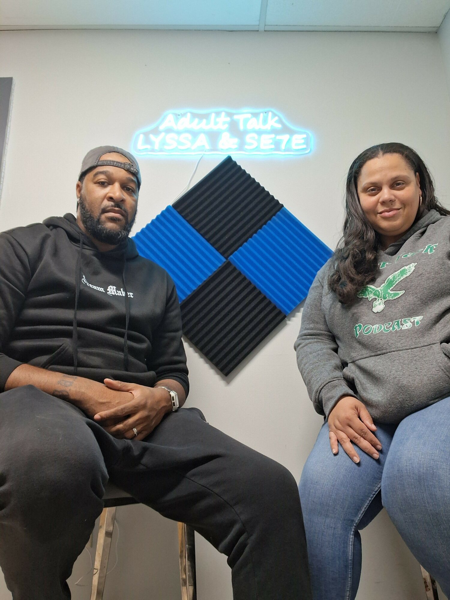 Two people sitting in a room with a neon sign and soundproof foam panels on the wall.