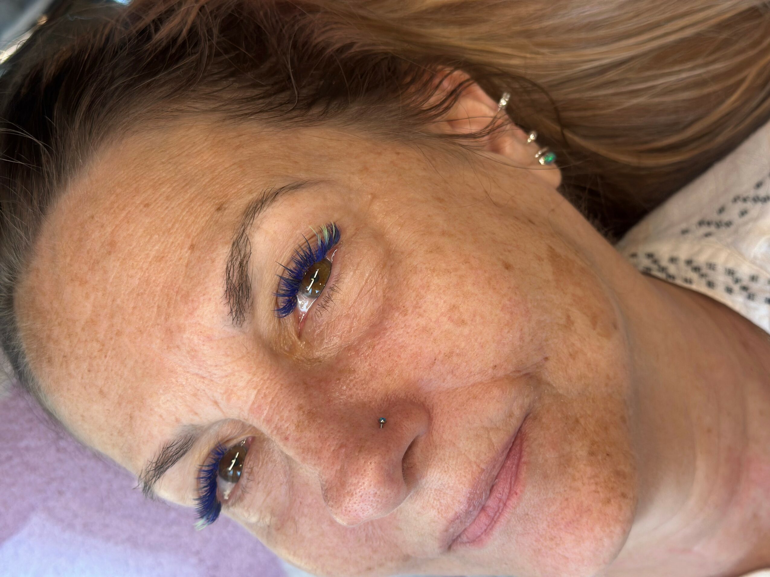 Close-up of a woman with freckles, lying down with eyes partially closed, wearing earrings and makeup, with a neutral expression.