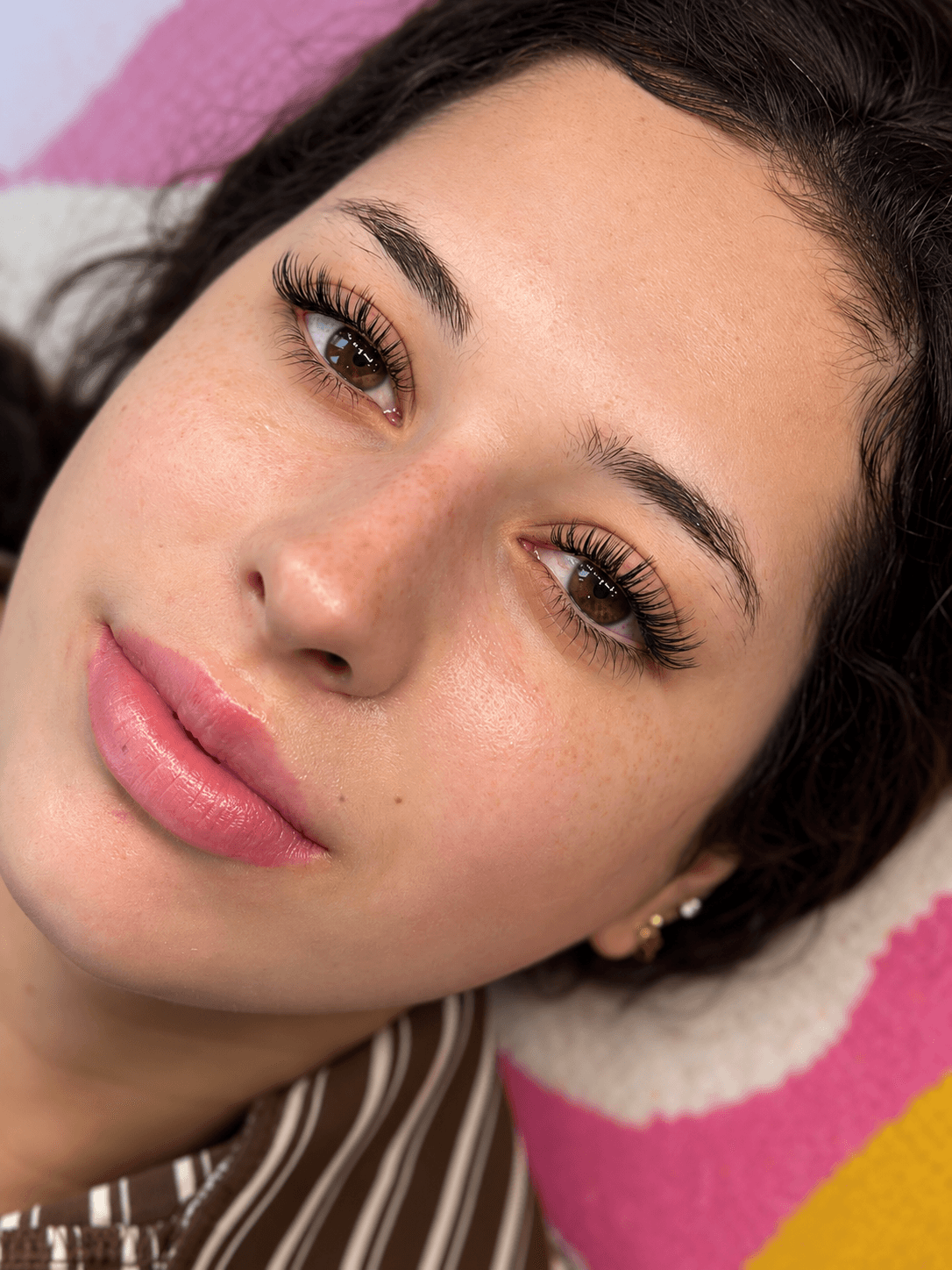 Close-up of a woman's face with makeup, lying on a pink and yellow blanket, looking at the camera.