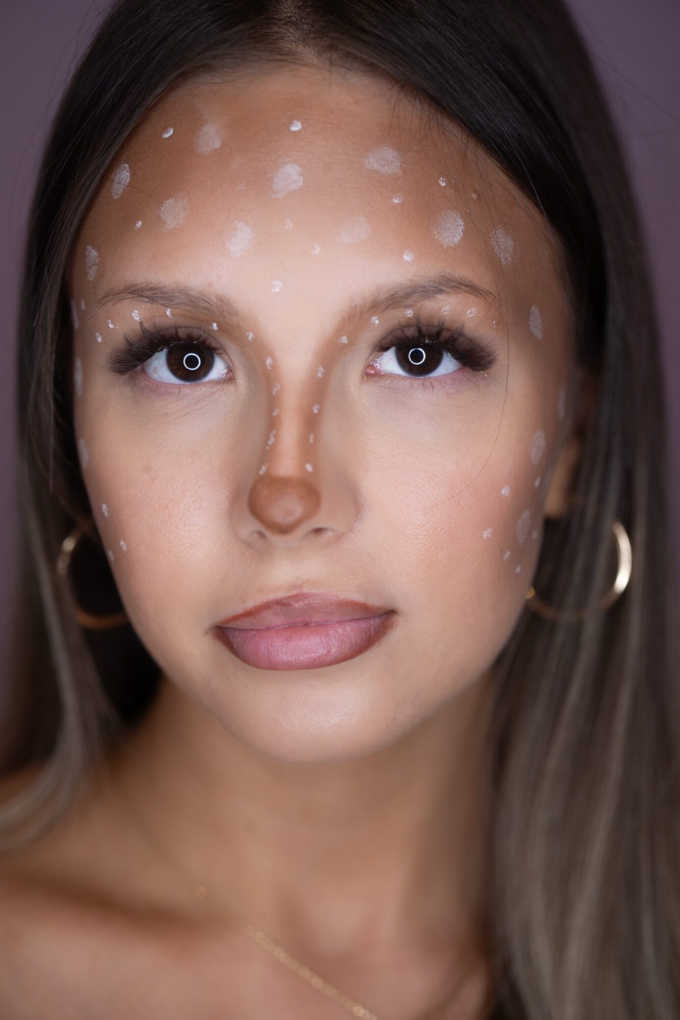 Close-up of a woman with dark hair, wearing hoop earrings, with white spots on her face, neutral expression, and makeup.