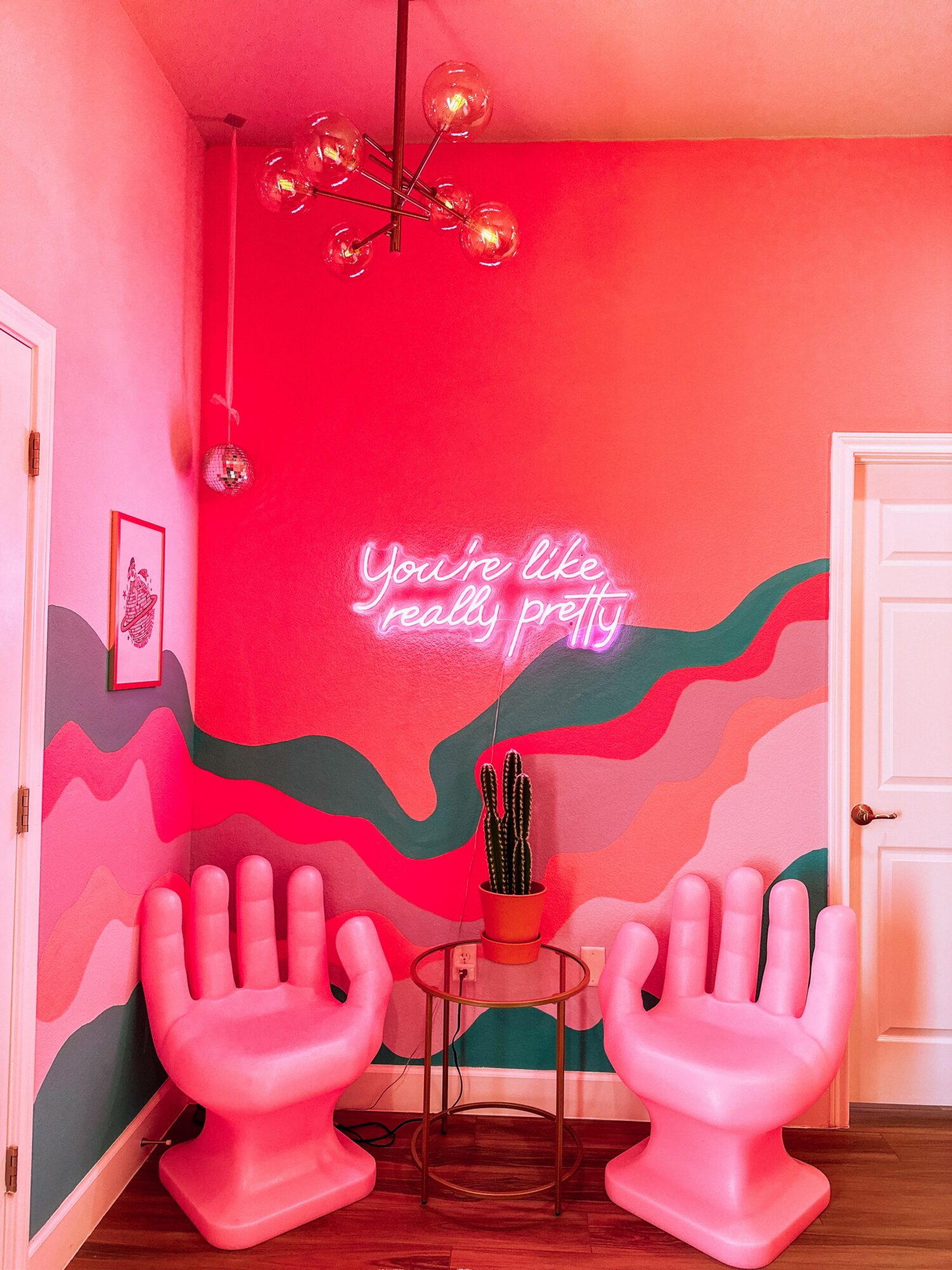 Pink room with a neon sign, two large hand-shaped chairs, and a cactus on a small table, colorful wall art, and a ceiling light.