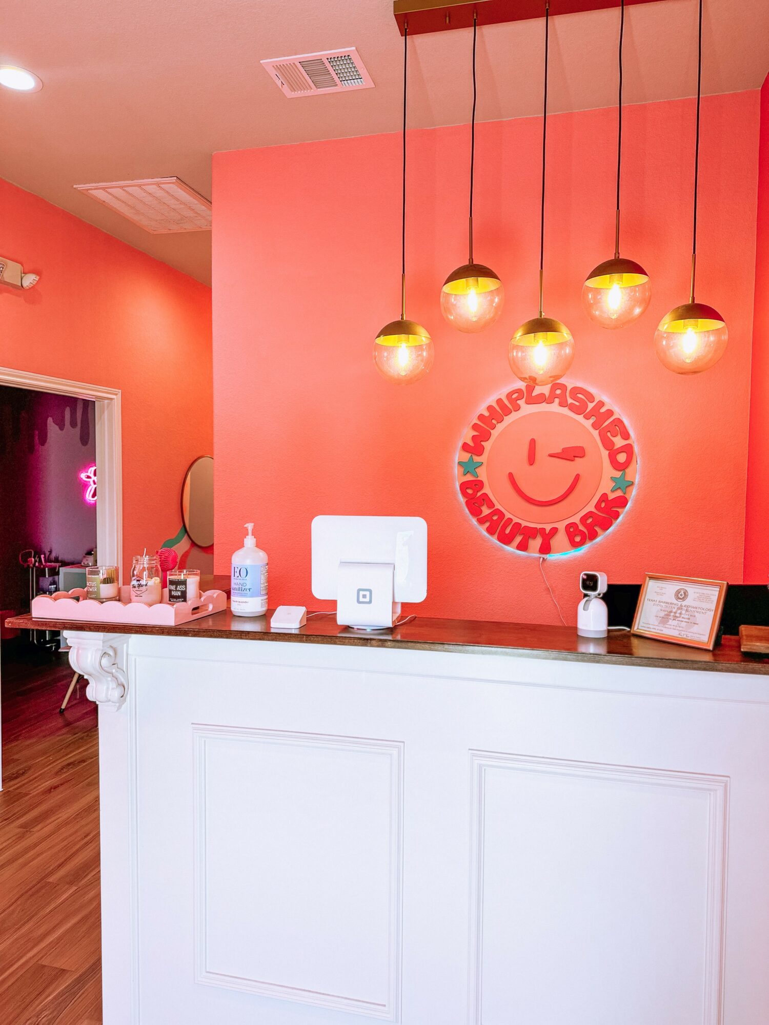Reception desk with sanitizer, a computer, and a sign, against a coral wall with a logo and hanging lights.