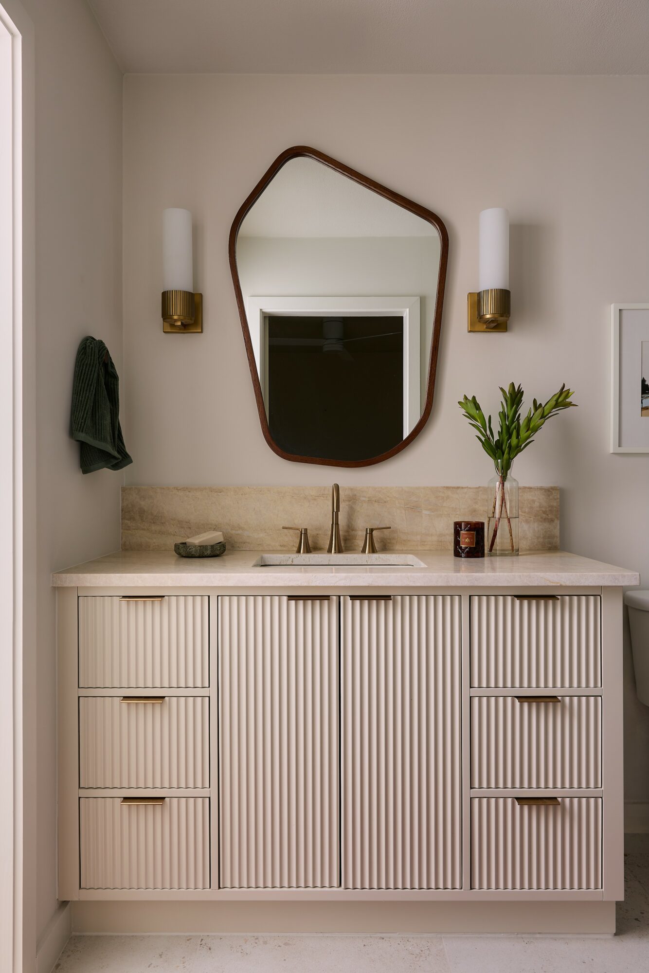 Bathroom vanity with striped cabinet, mirror, two wall sconces, and a plant on the right, with a towel on the left.
